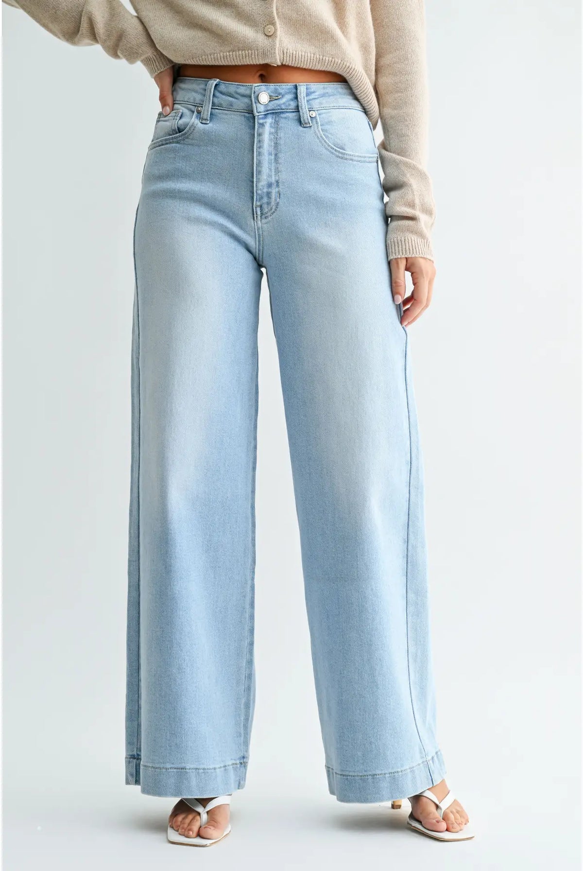 A person wearing light-wash wide-leg denim jeans with a beige cardigan and white sandals, shown from the waist down against a plain light gray studio background.