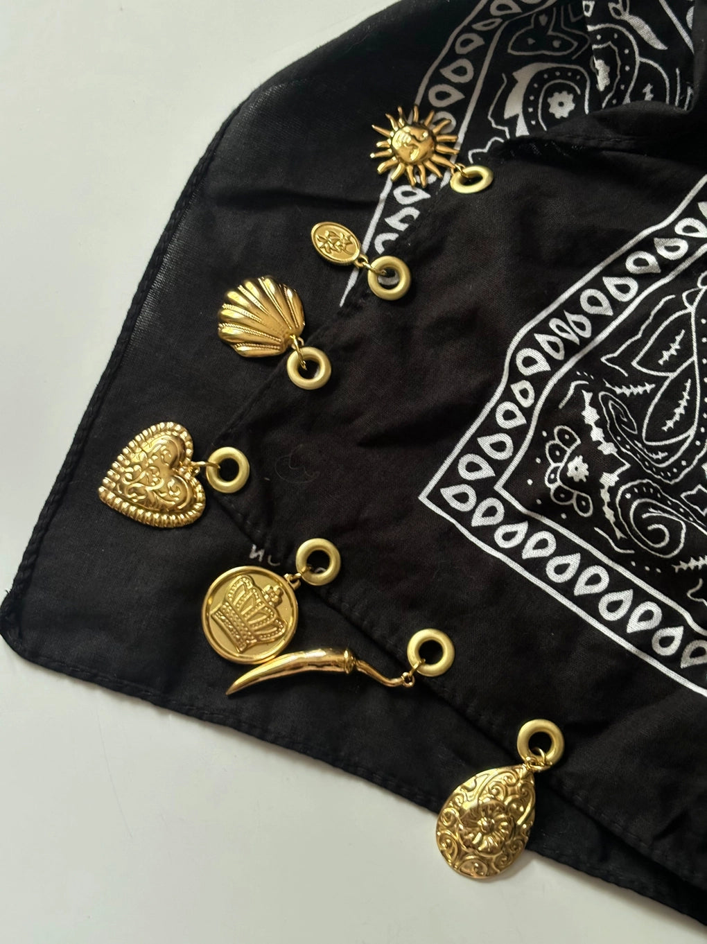 A black bandana scarf with a white paisley print, styled with multiple gold-toned metal charms including a sun, shell, heart, coin, crescent moon, and teardrop pendant.