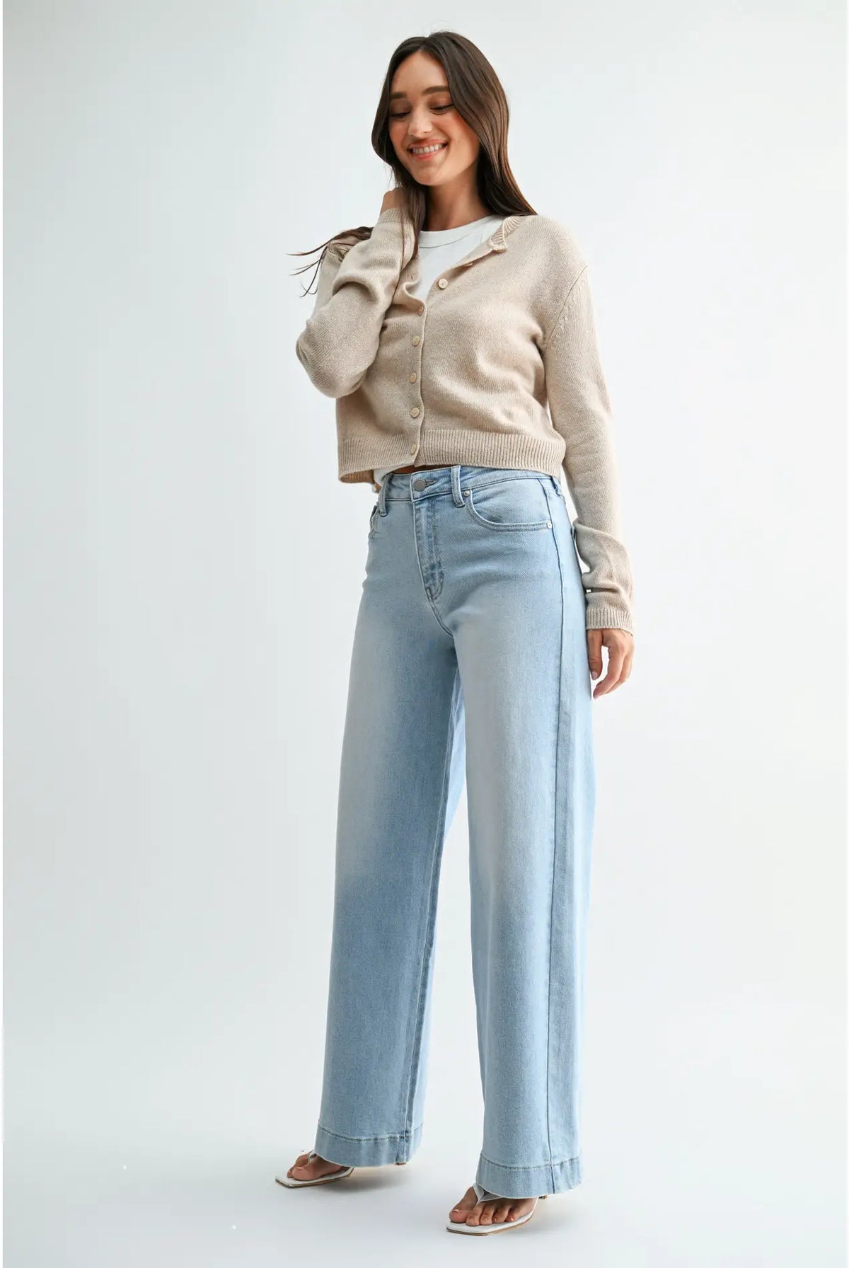 A woman modeling a light beige cropped cardigan over a white top and light blue wide-leg jeans, standing against a plain light gray studio background.
