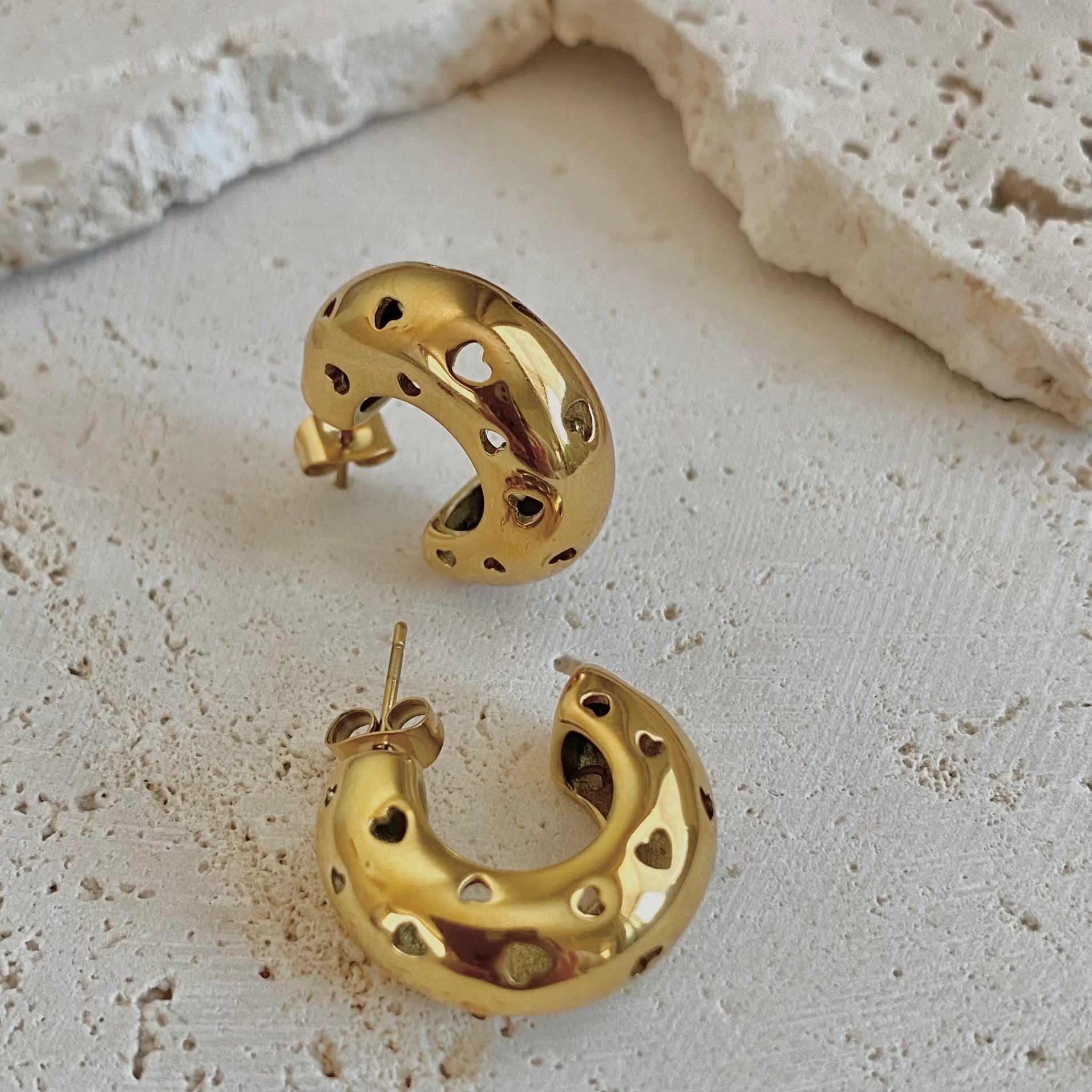 Gold crescent-shaped hoop earrings with heart-shaped cutouts, displayed on a textured light beige stone surface.