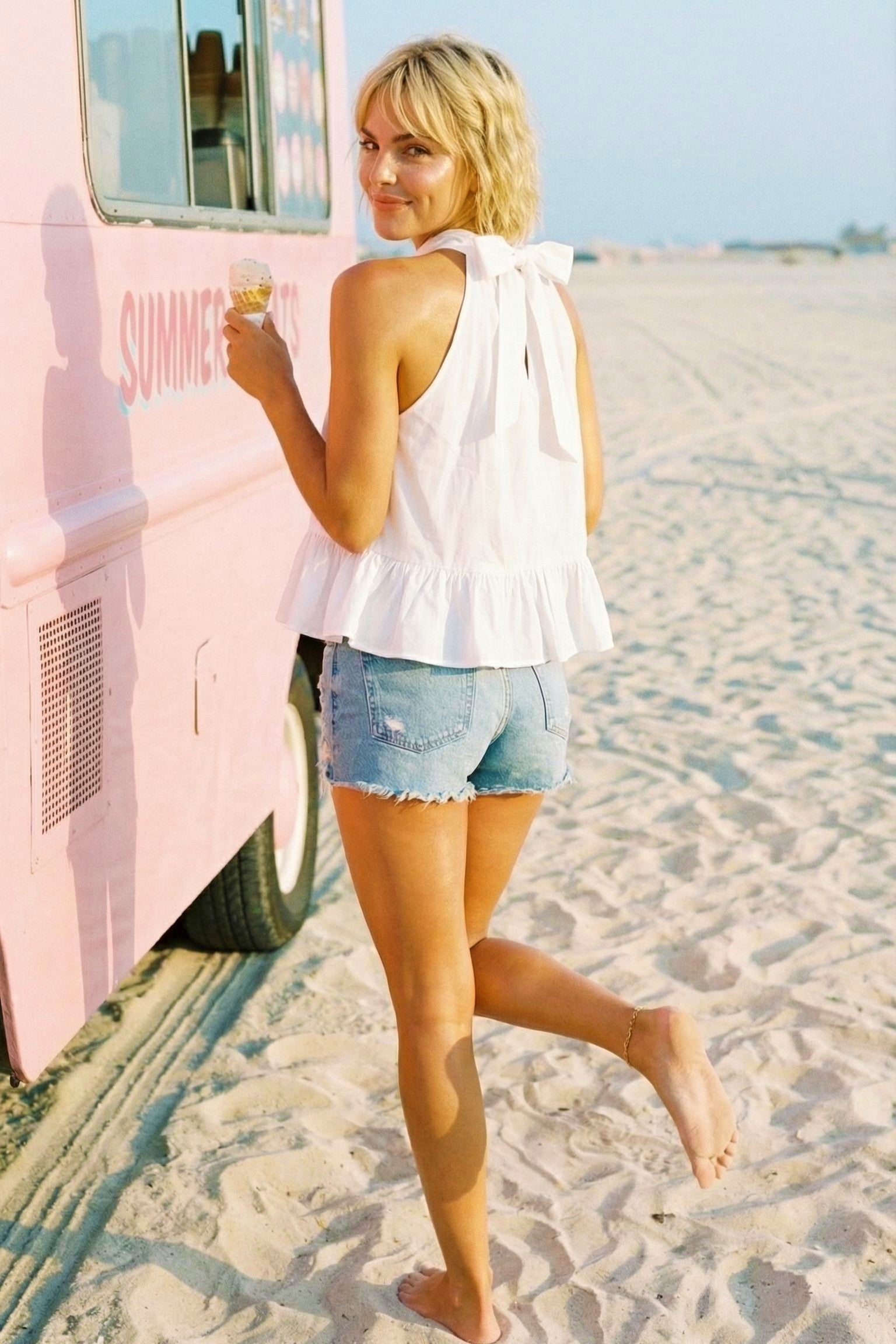 Woman in a white top and denim shorts standing on a sandy beach next to a pink vehicle.