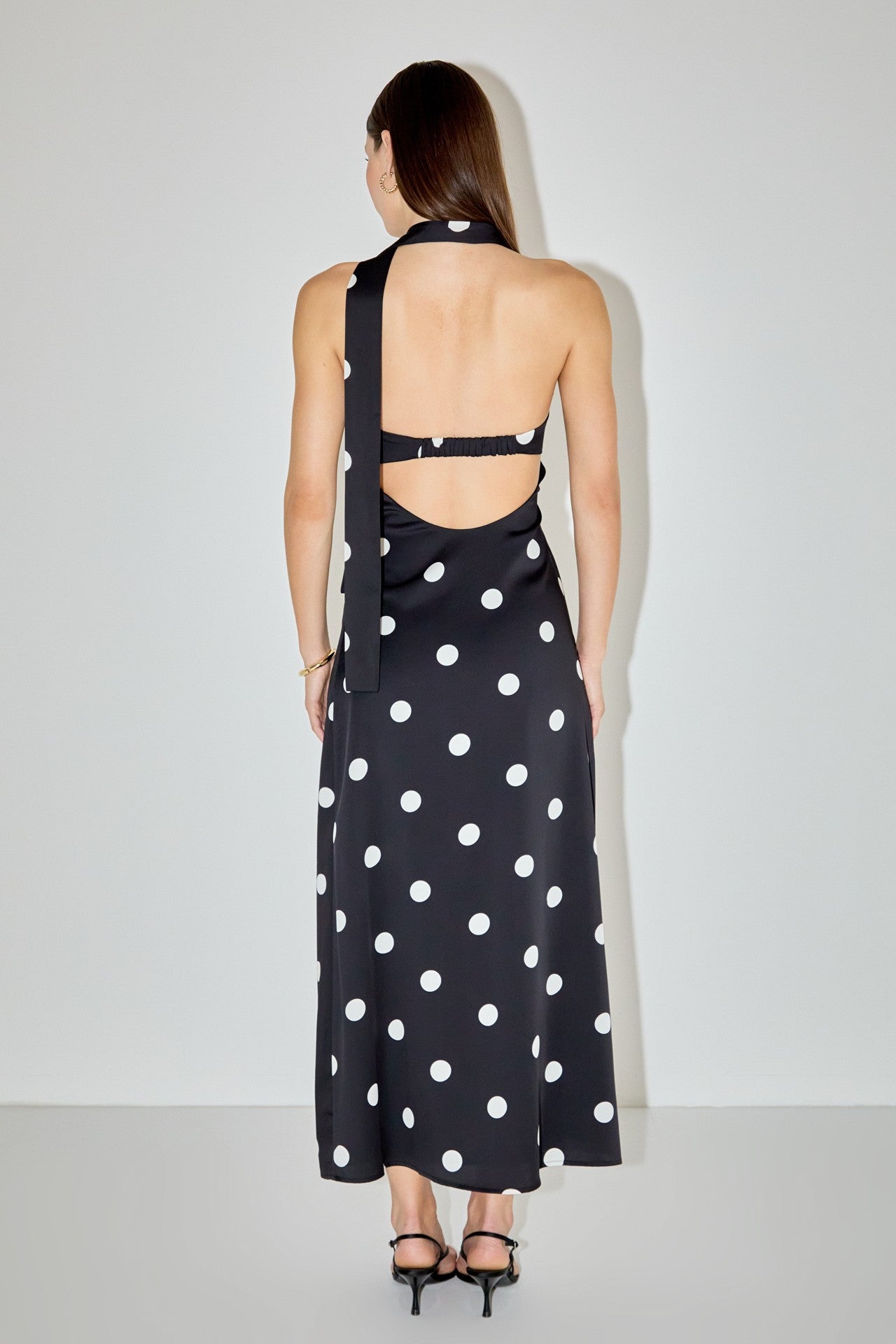 A woman viewed from the back wearing a black maxi dress with a white polka dot pattern and an open back cutout, paired with black strappy heels against a plain light gray studio background.
