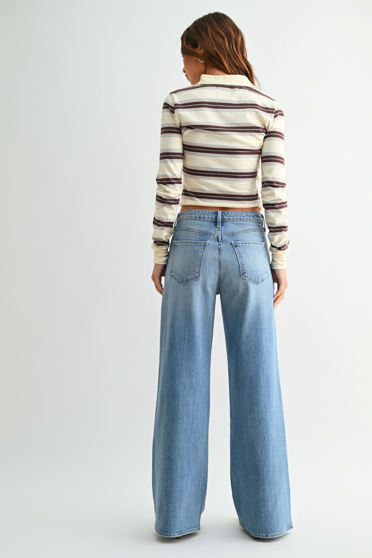 A woman shown from the back wearing a cream long-sleeve striped top with brown and black stripes, paired with light-wash wide-leg blue jeans, against a plain light gray studio background.