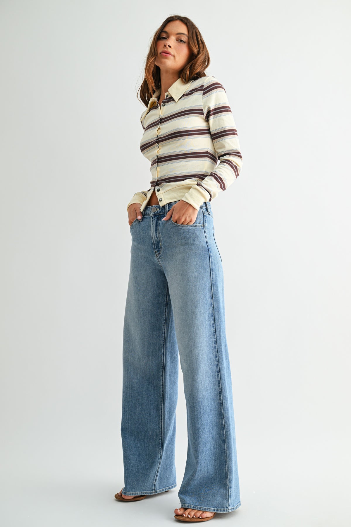 A woman modeling a cream long-sleeve knit polo shirt with brown and black horizontal stripes, paired with light-wash wide-leg blue jeans and brown flat sandals, against a light gray studio background.