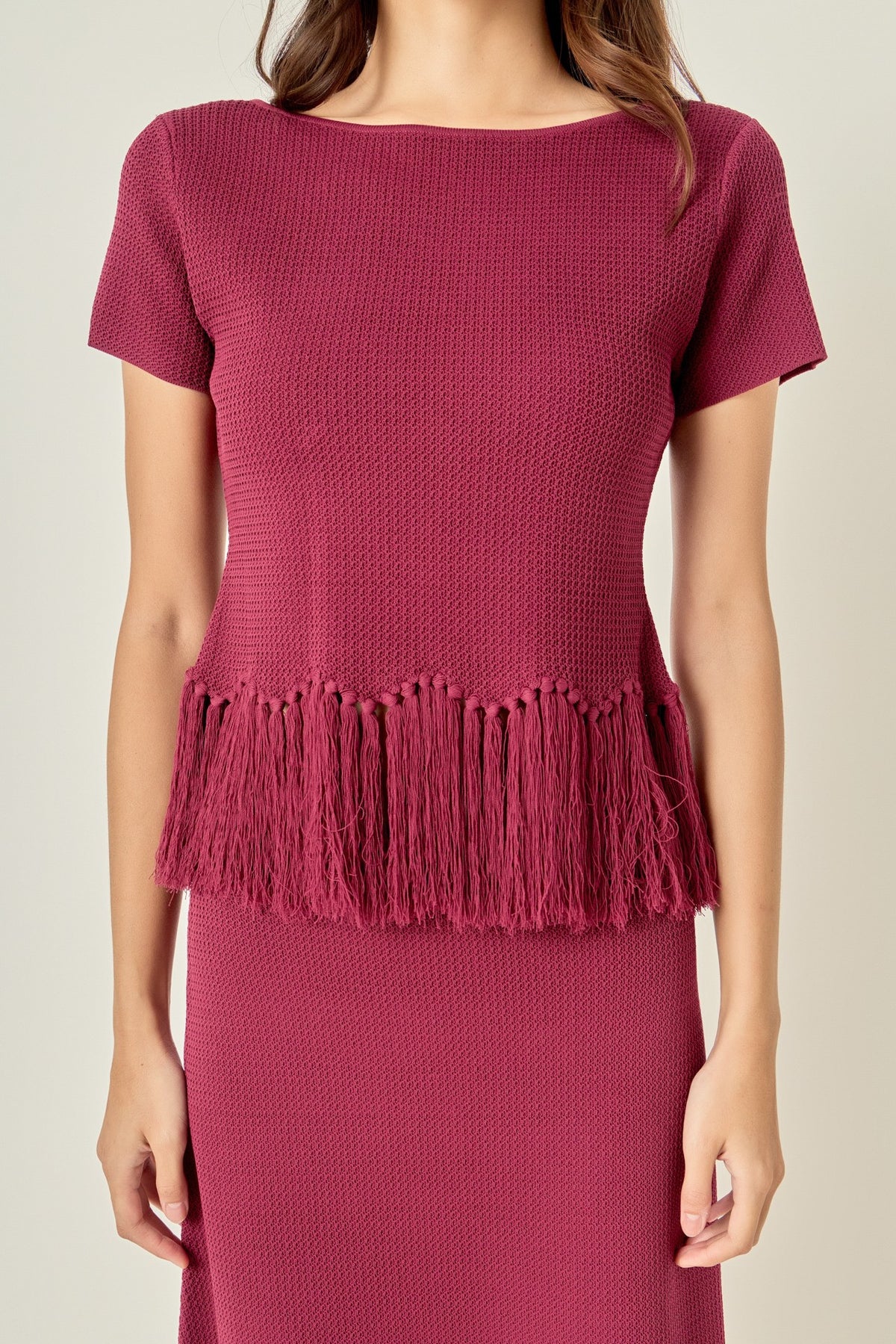 A woman wearing a textured short-sleeve top in a deep berry pink, featuring a fringed hem and a matching textured skirt against a light beige background.