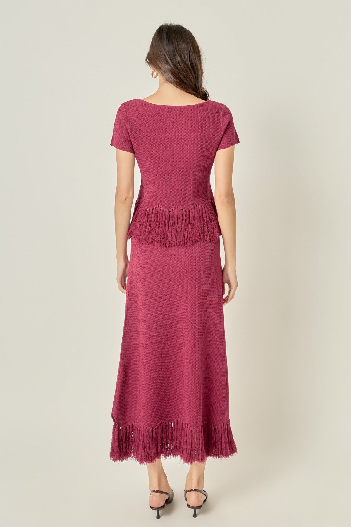 A woman viewed from the back wearing a magenta maxi dress with short sleeves, a crew neckline, and fringe detailing at the waist and hem, paired with black high-heeled sandals against a light beige studio background.