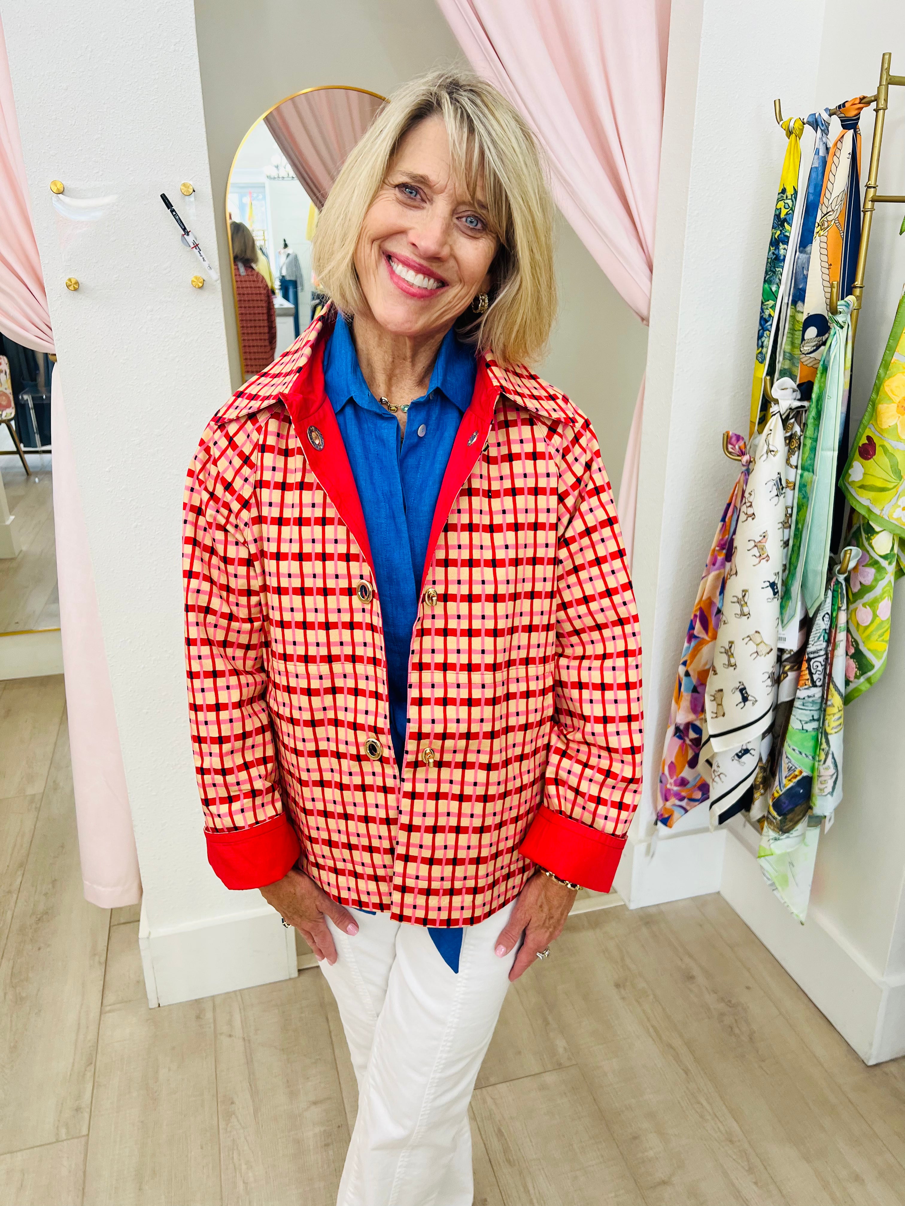 A smiling blonde woman stands in a boutique wearing a red and white patterned blazer over a bright blue collared shirt with white pants. Pink curtains, a mirror, and a clothing rack are visible in the background.
