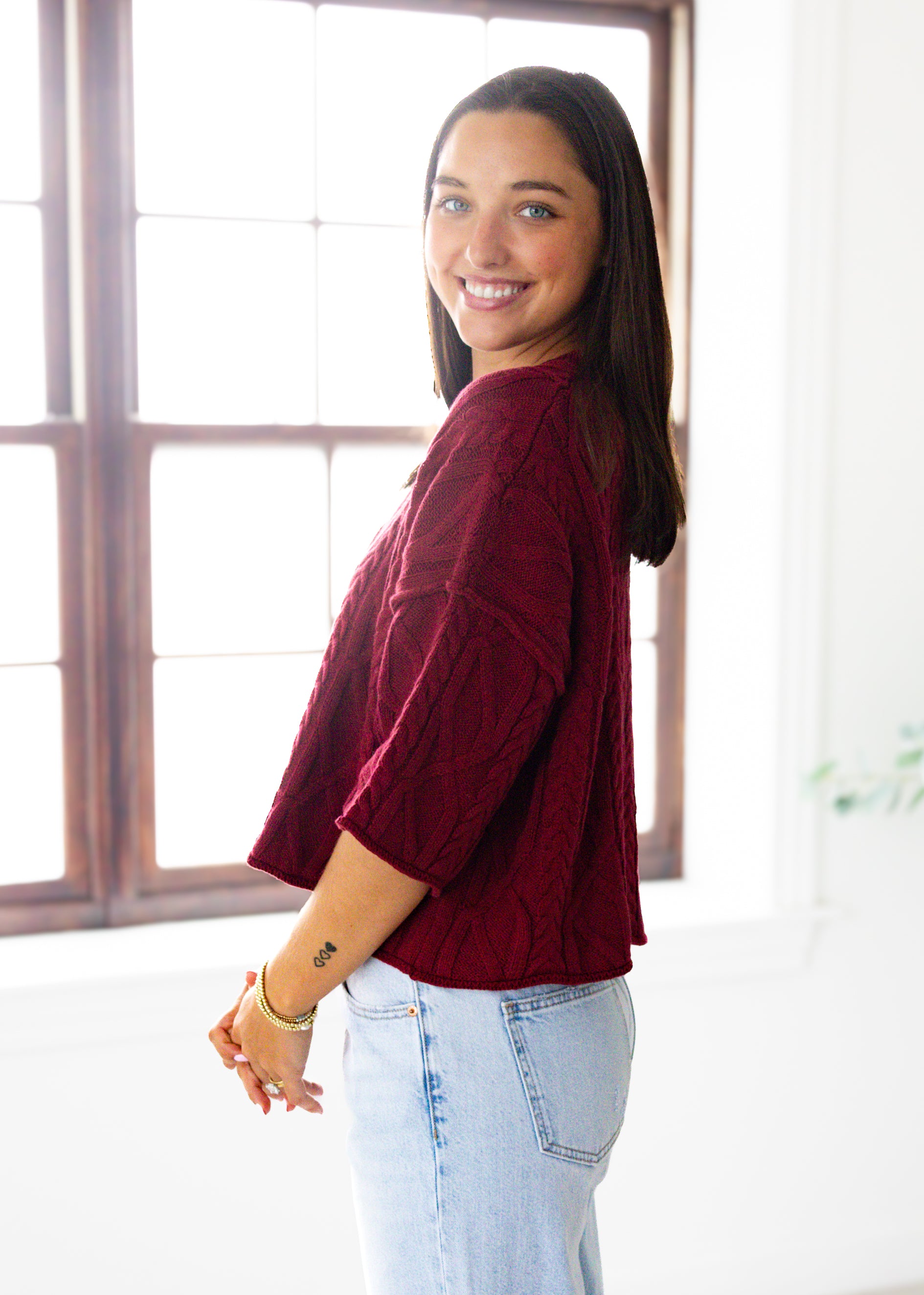 Woman wearing a burgundy sweater and light blue jeans standing in a room with large windows.