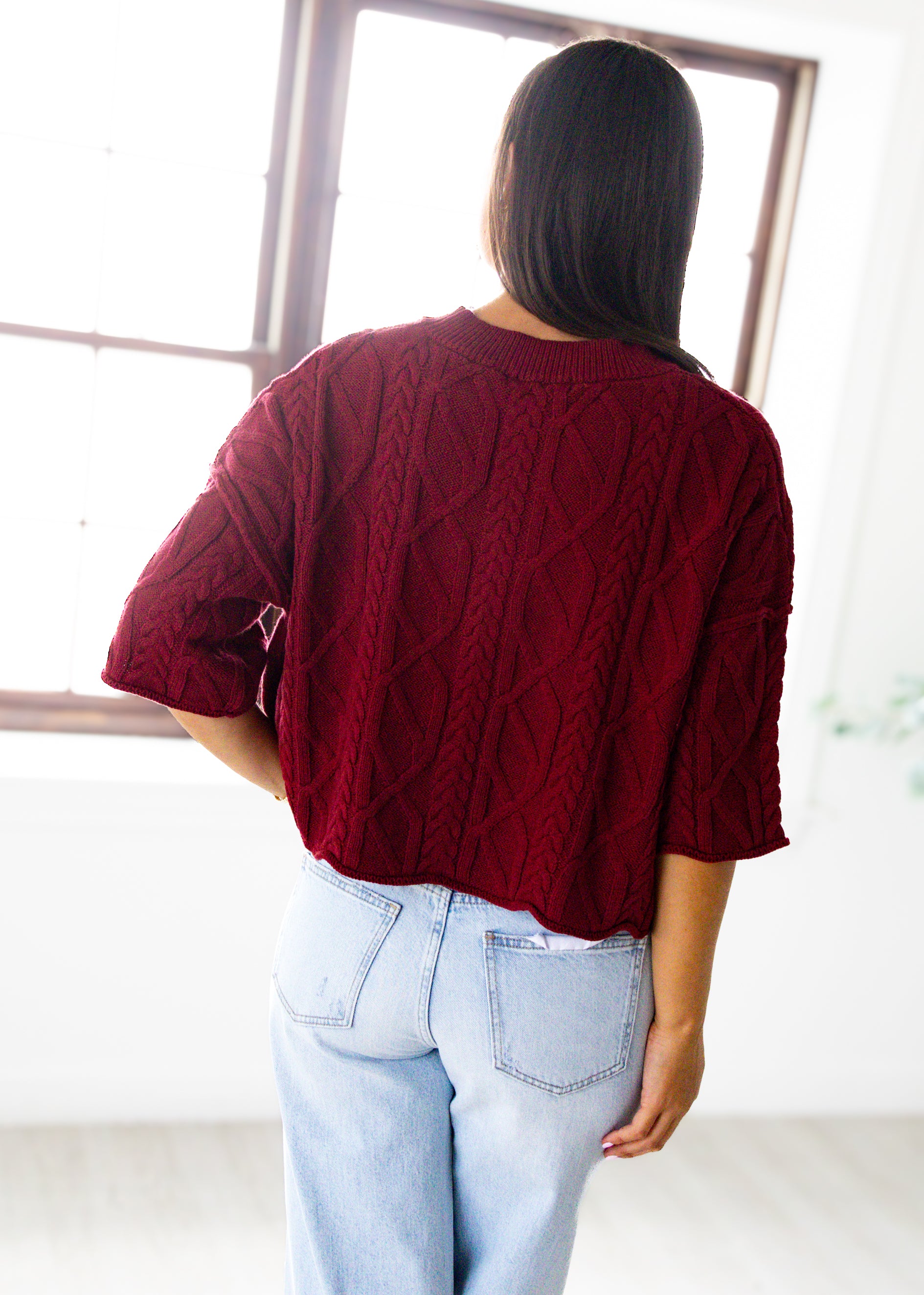 Person wearing a burgundy cable knit sweater and light blue jeans, standing in front of a mirror.