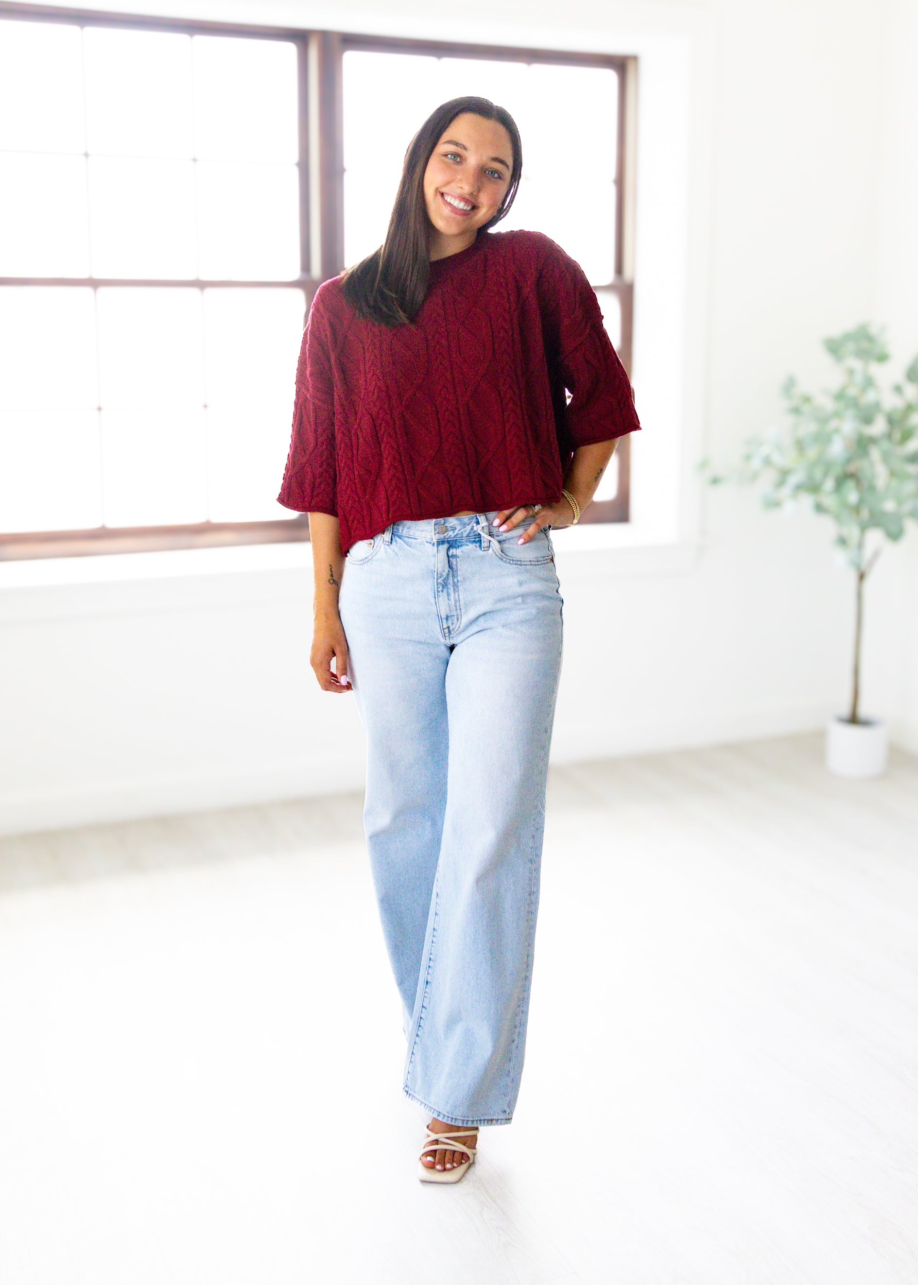 Woman wearing a burgundy sweater and light blue jeans standing in a bright room with a plant in the background.
