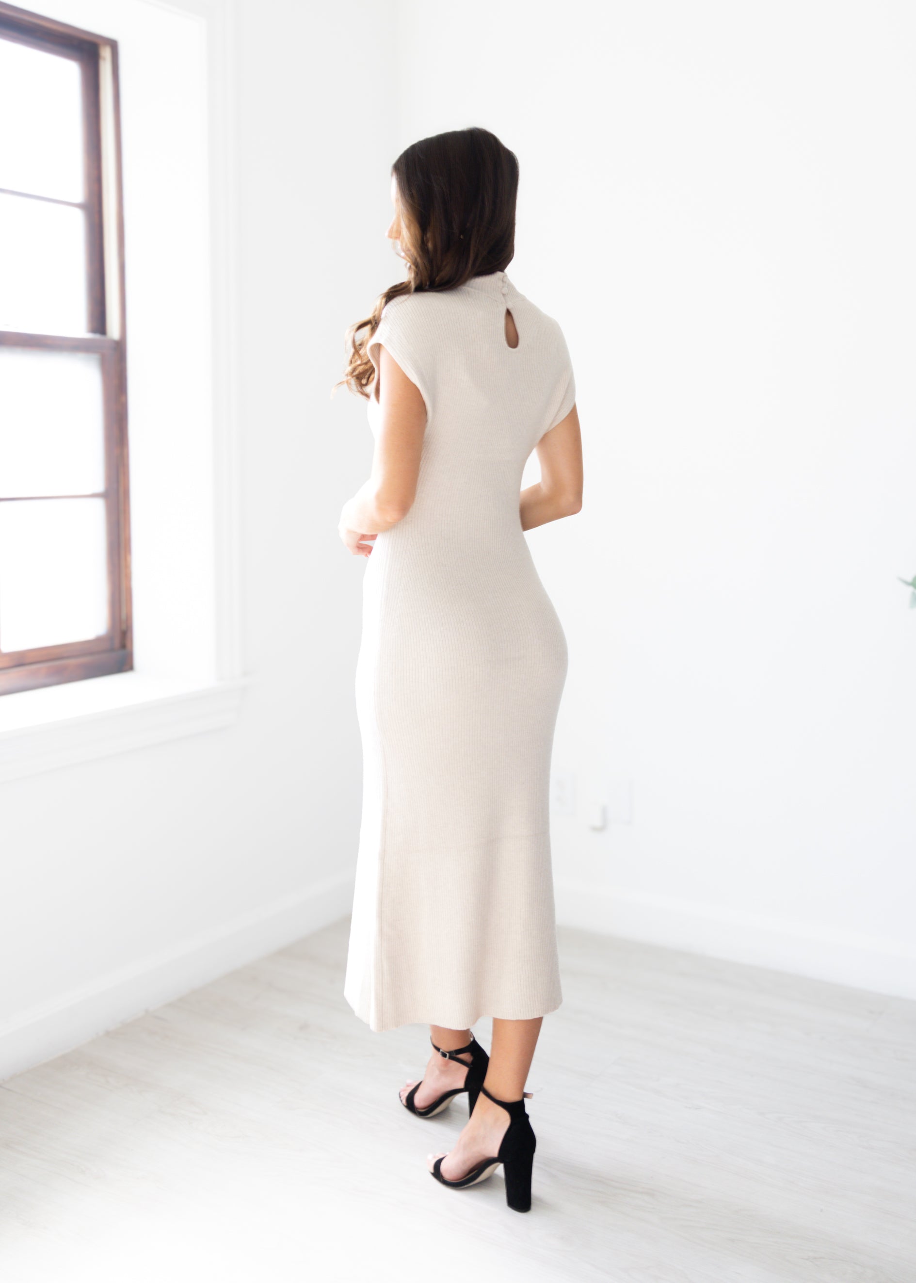Woman wearing a tan dress standing in a minimalistic room with a window.