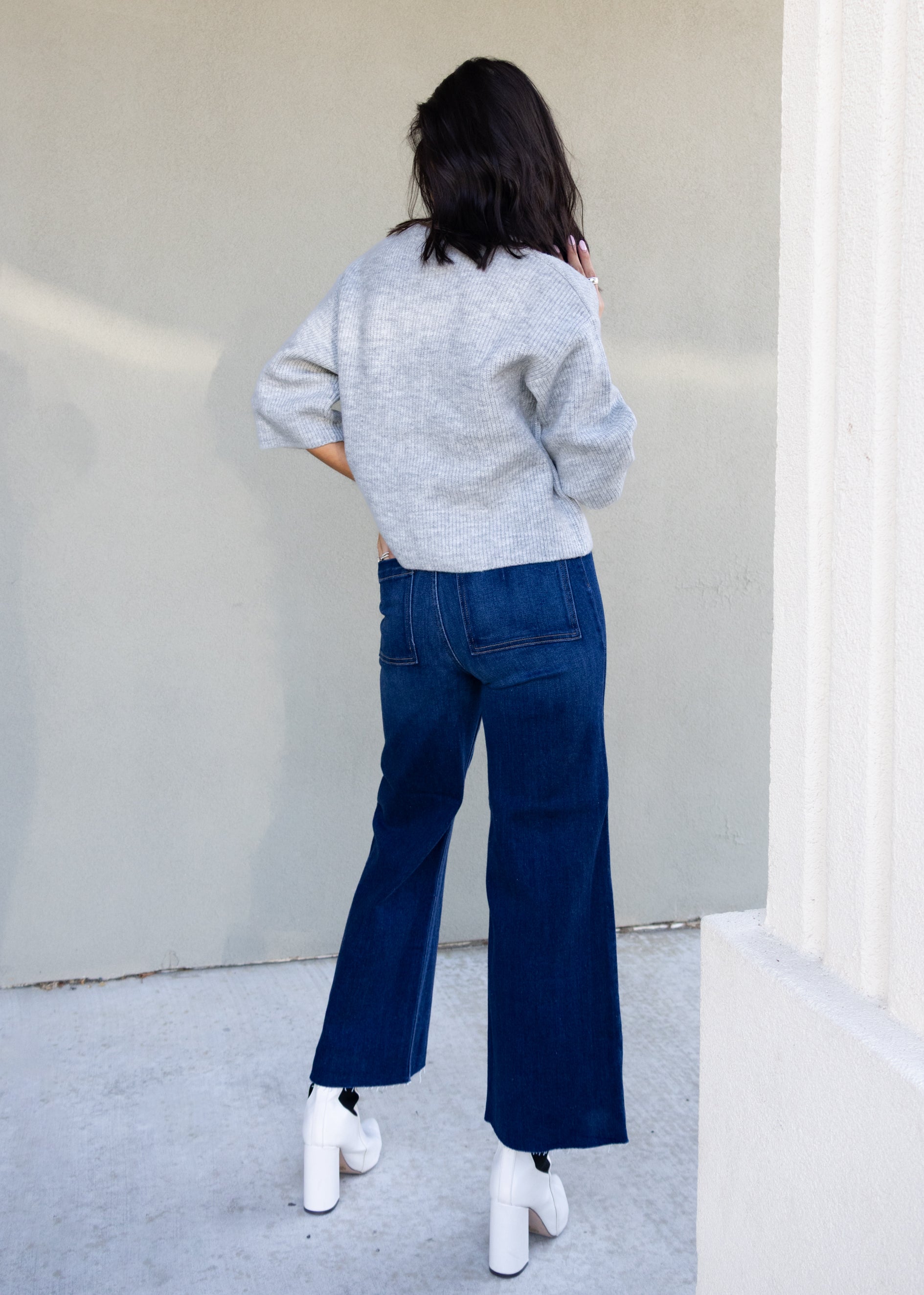 Person wearing a gray sweater and blue jeans standing against a white wall.