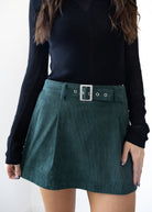 Person wearing a dark green skirt with a belt and black long-sleeve top on a white background