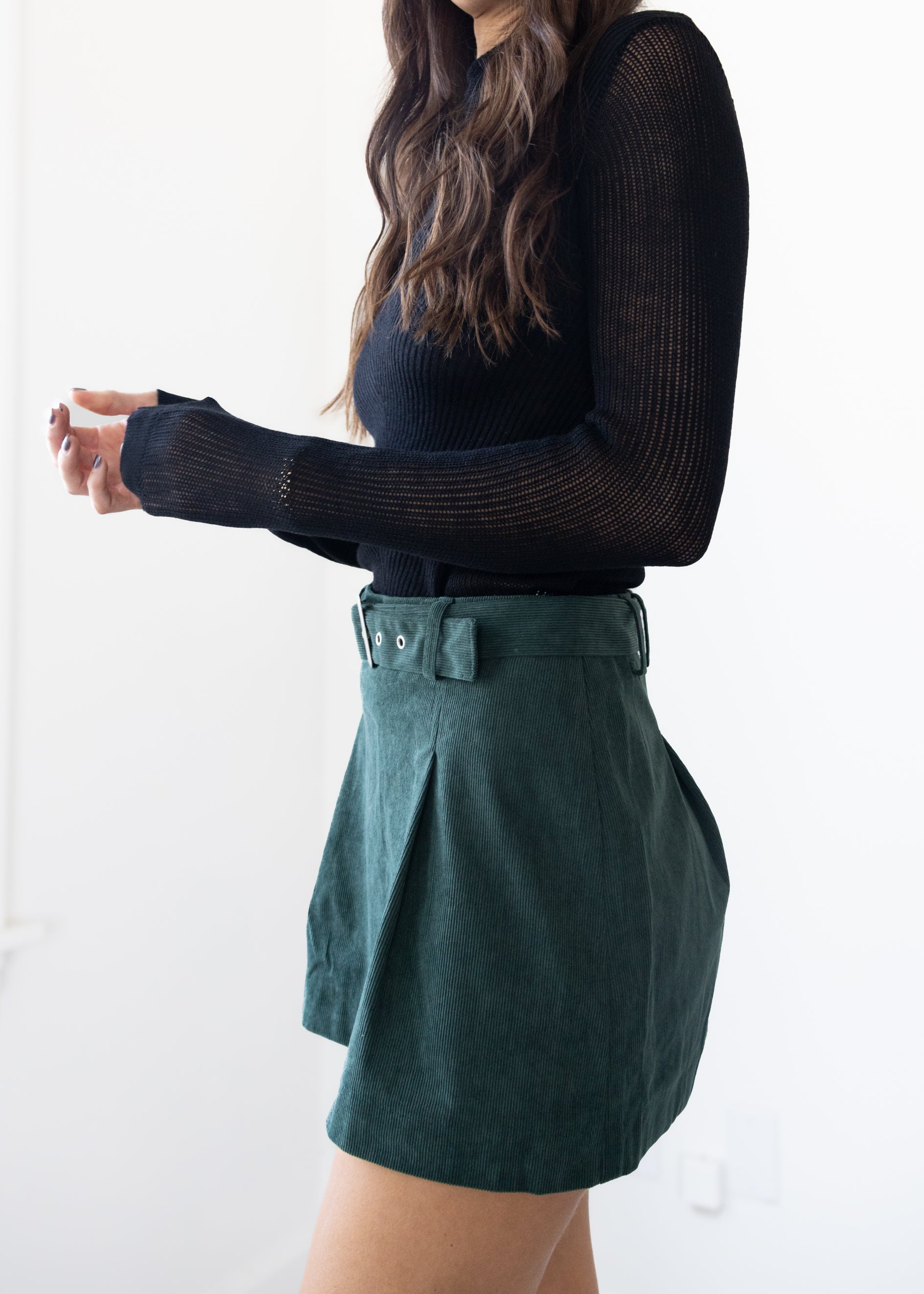Person wearing a black long-sleeve top and green pleated skirt on a white background
