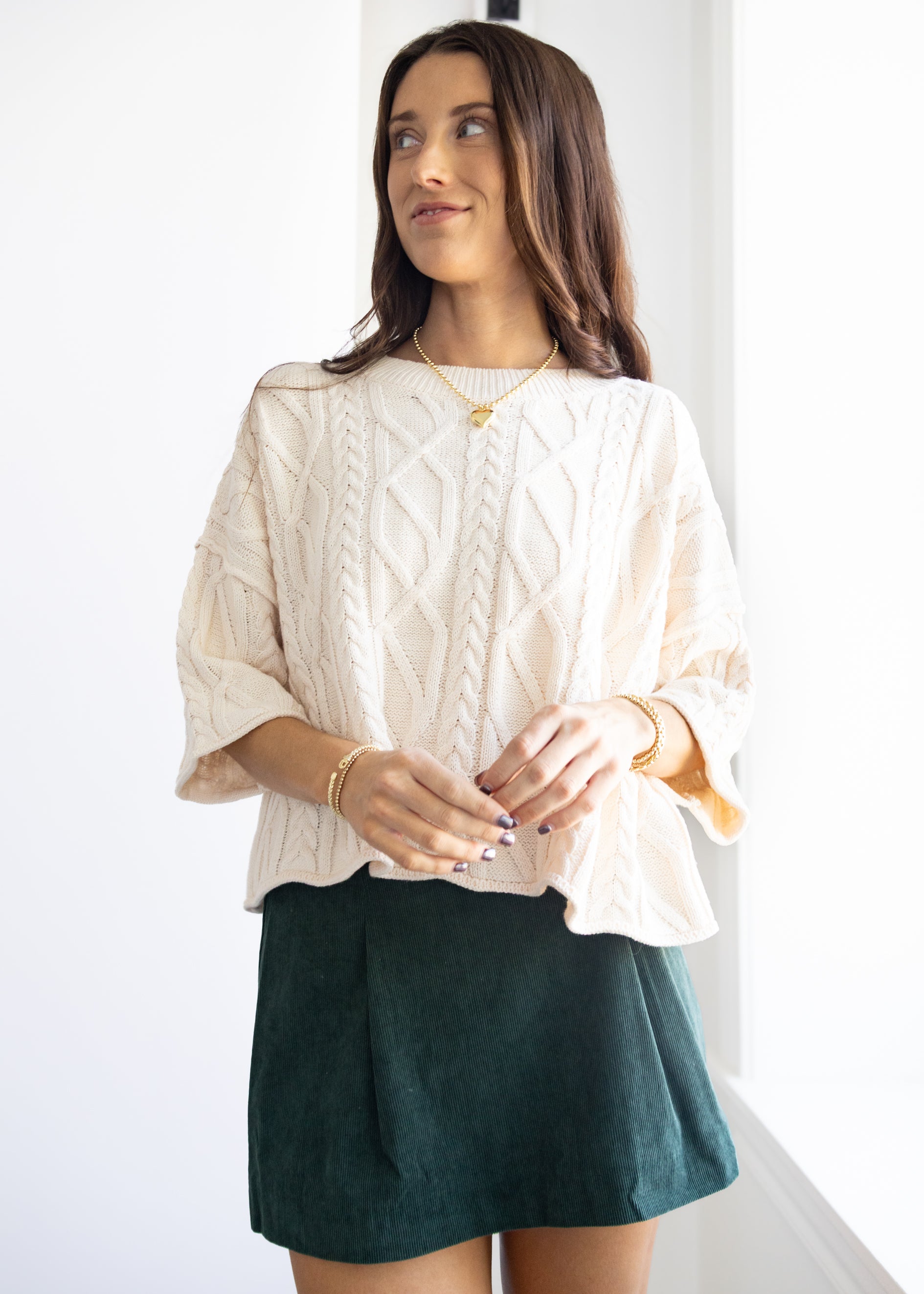 Woman wearing a white cable knit sweater and green skirt indoors.