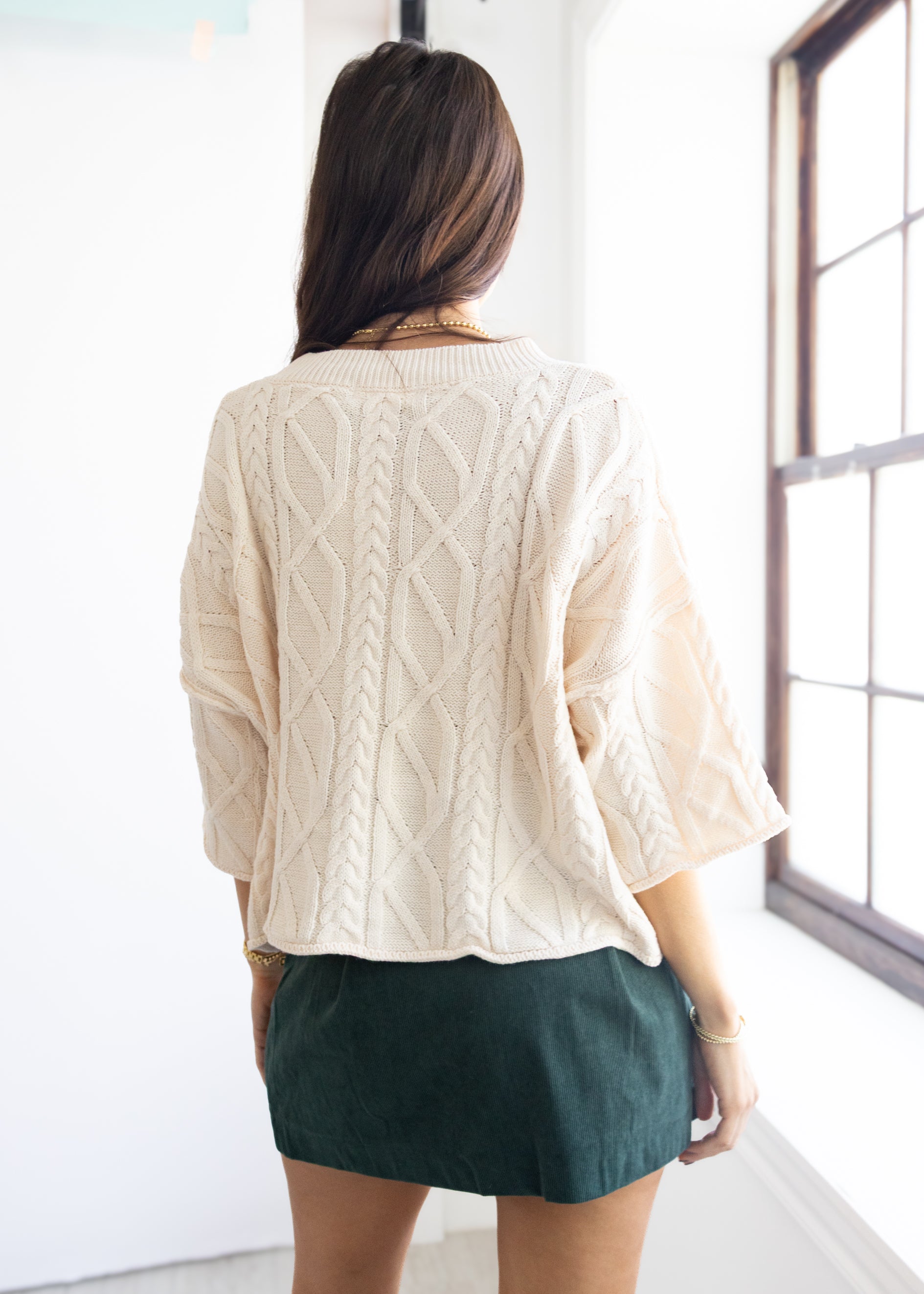 Person wearing a cream cable knit sweater and green skirt in a bright room.