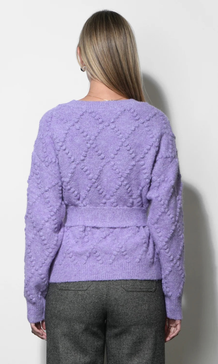 A woman viewed from the back wearing a lavender purple textured knit sweater with a waist tie and gray trousers, standing against a plain light gray wall.