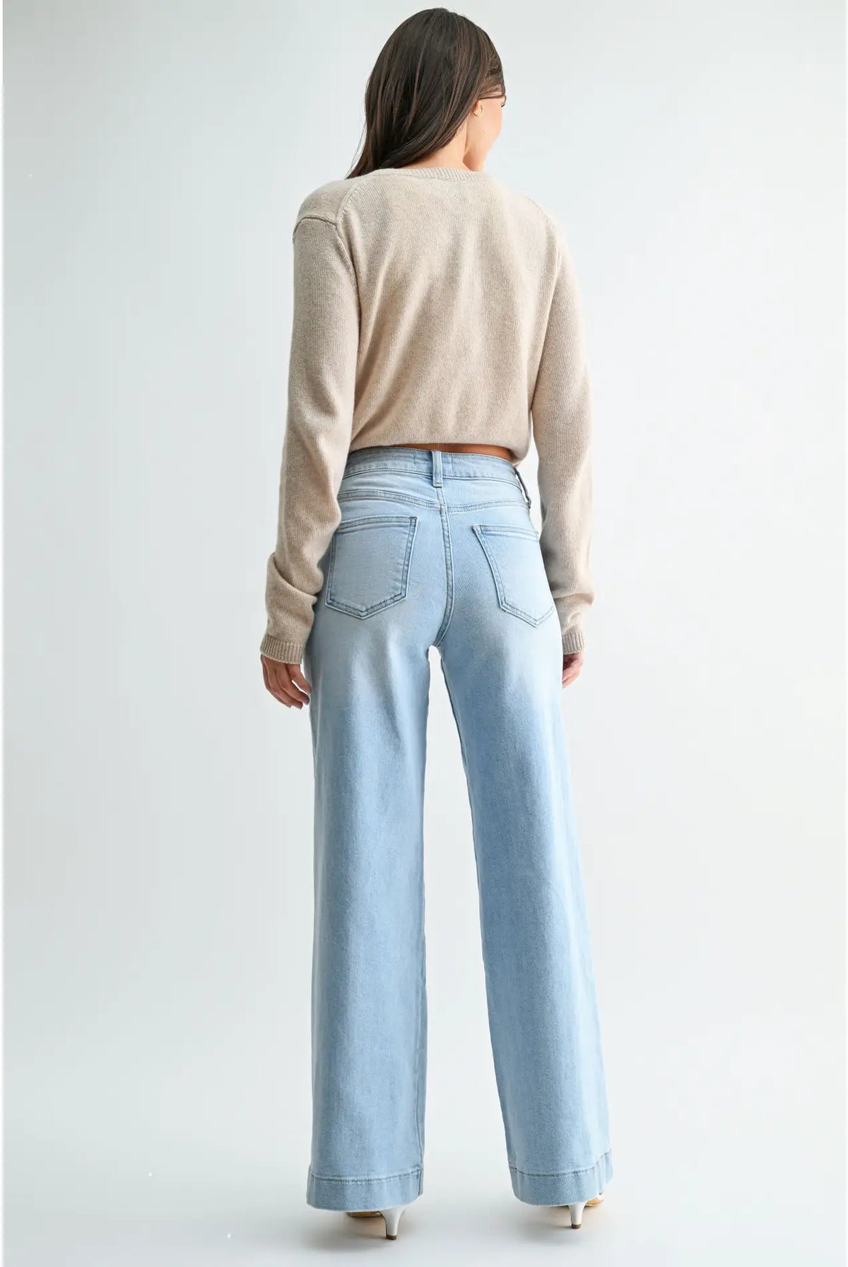 A woman shown from the back wearing a beige long-sleeve cropped sweater and light blue wide-leg jeans against a plain light gray studio background.