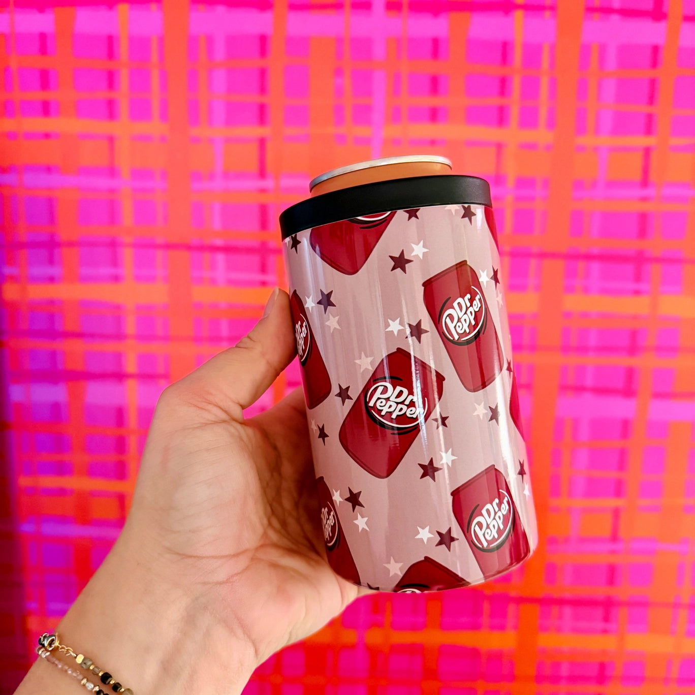Can cooler with Dr Pepper design held by a hand against a colorful plaid background