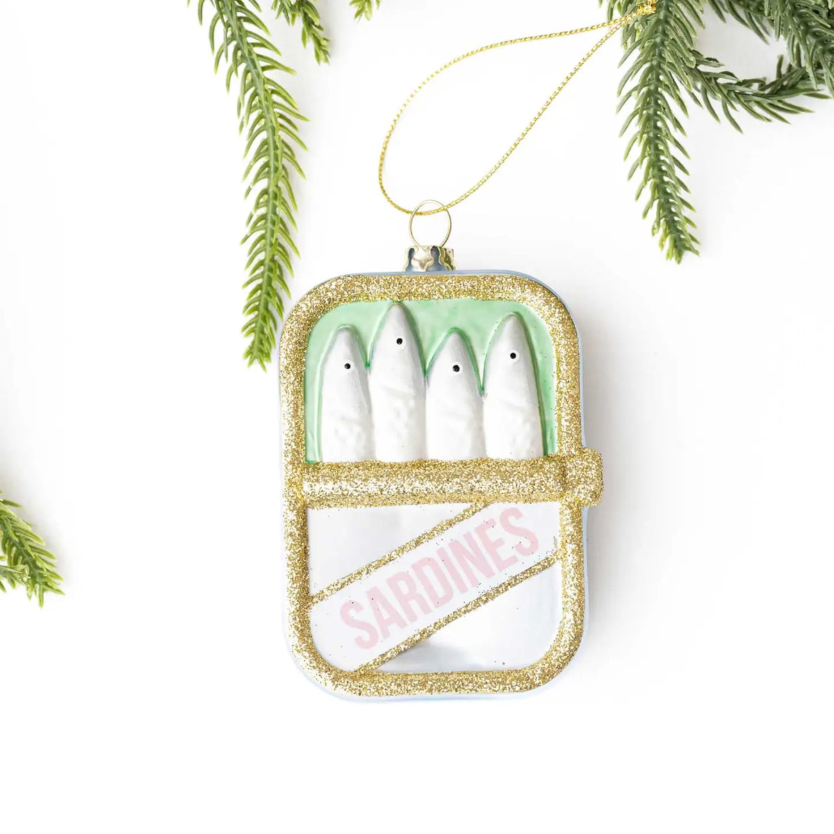 Decorative ornament shaped like a can of sardines with gold glitter and green accents, hanging against a white background with greenery.