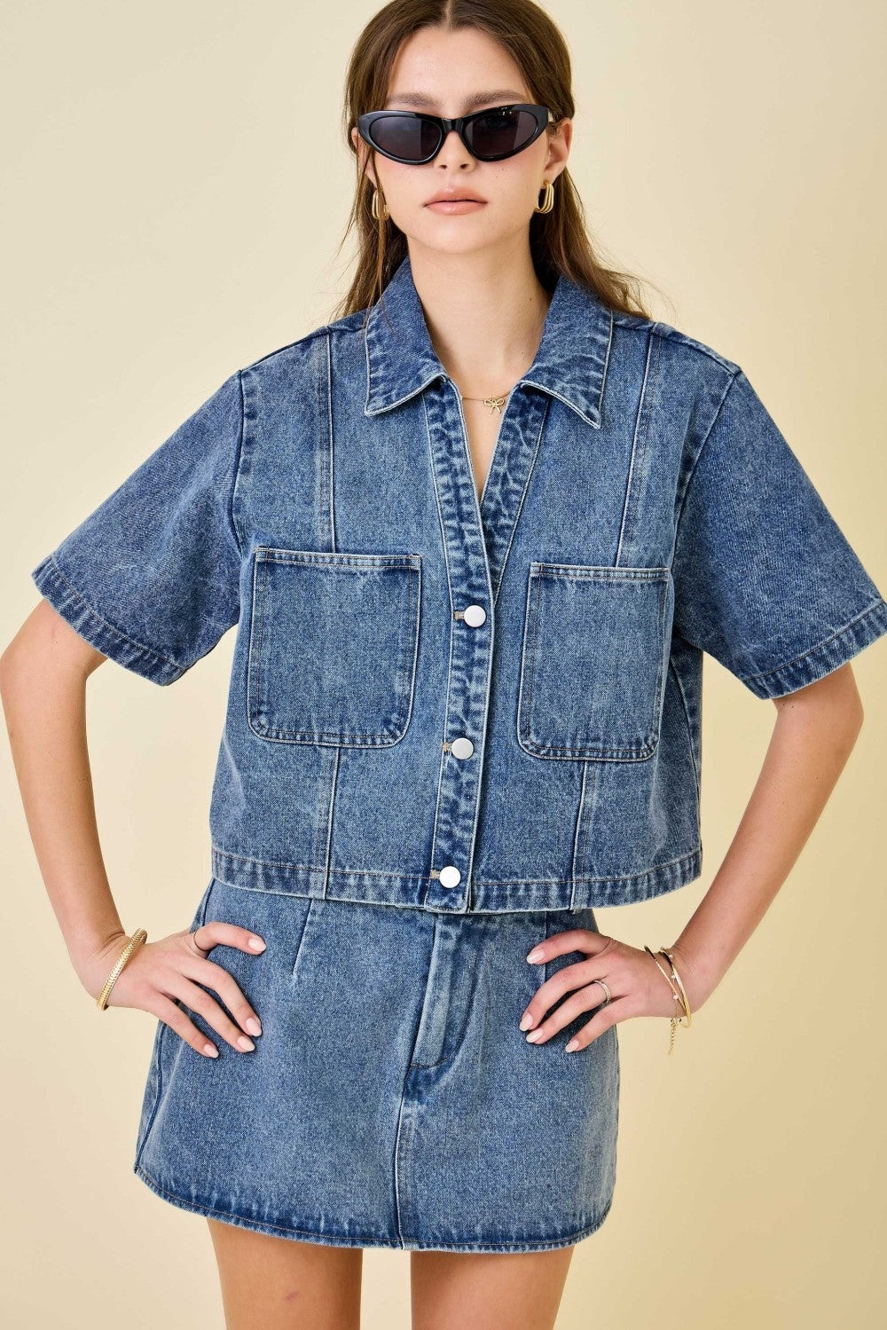 Woman wearing a denim jacket and skirt set with sunglasses on a beige background
