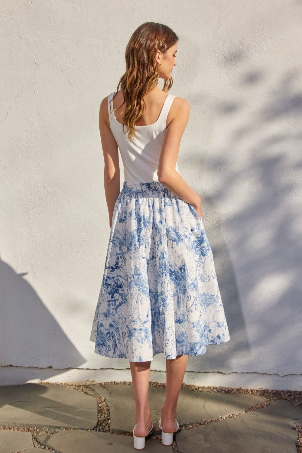 Woman wearing a white tank top and blue floral skirt against a light wall.
