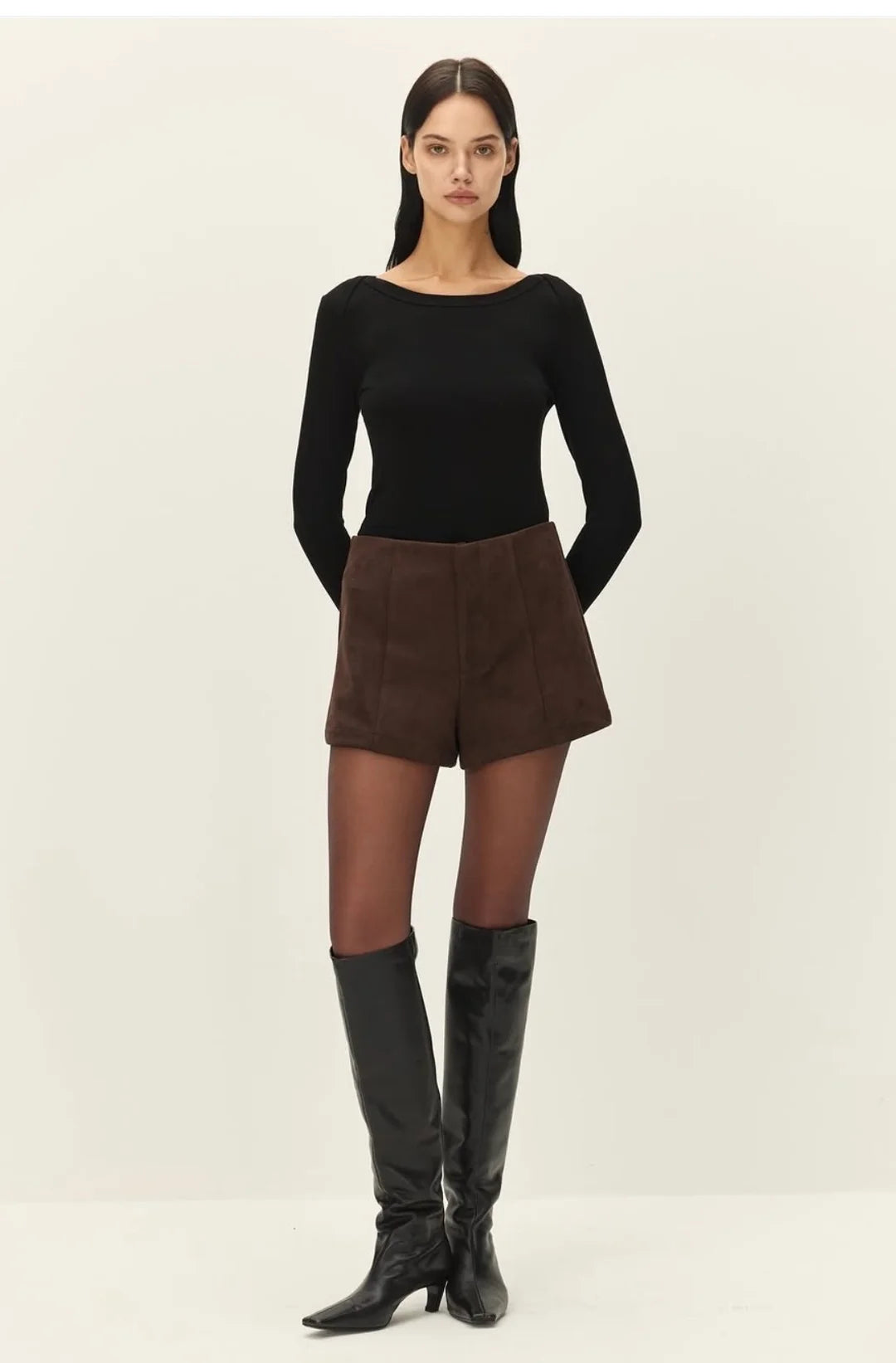 A model stands against a plain light background wearing a fitted black long-sleeve top, brown shorts, sheer black tights, and black knee-high boots.