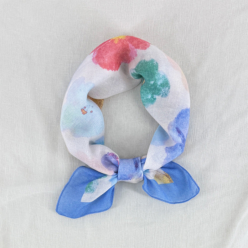 A lightweight silk scarf with a watercolor floral print, tied in a loop with a bow at the bottom, displayed on a textured off-white fabric background.