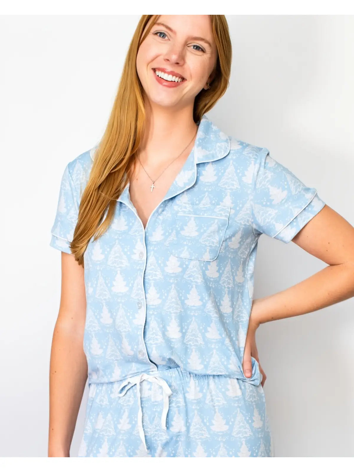 A woman wearing a light blue short-sleeve pajama top with a white pine tree pattern, paired with matching drawstring bottoms, against a plain light gray background.