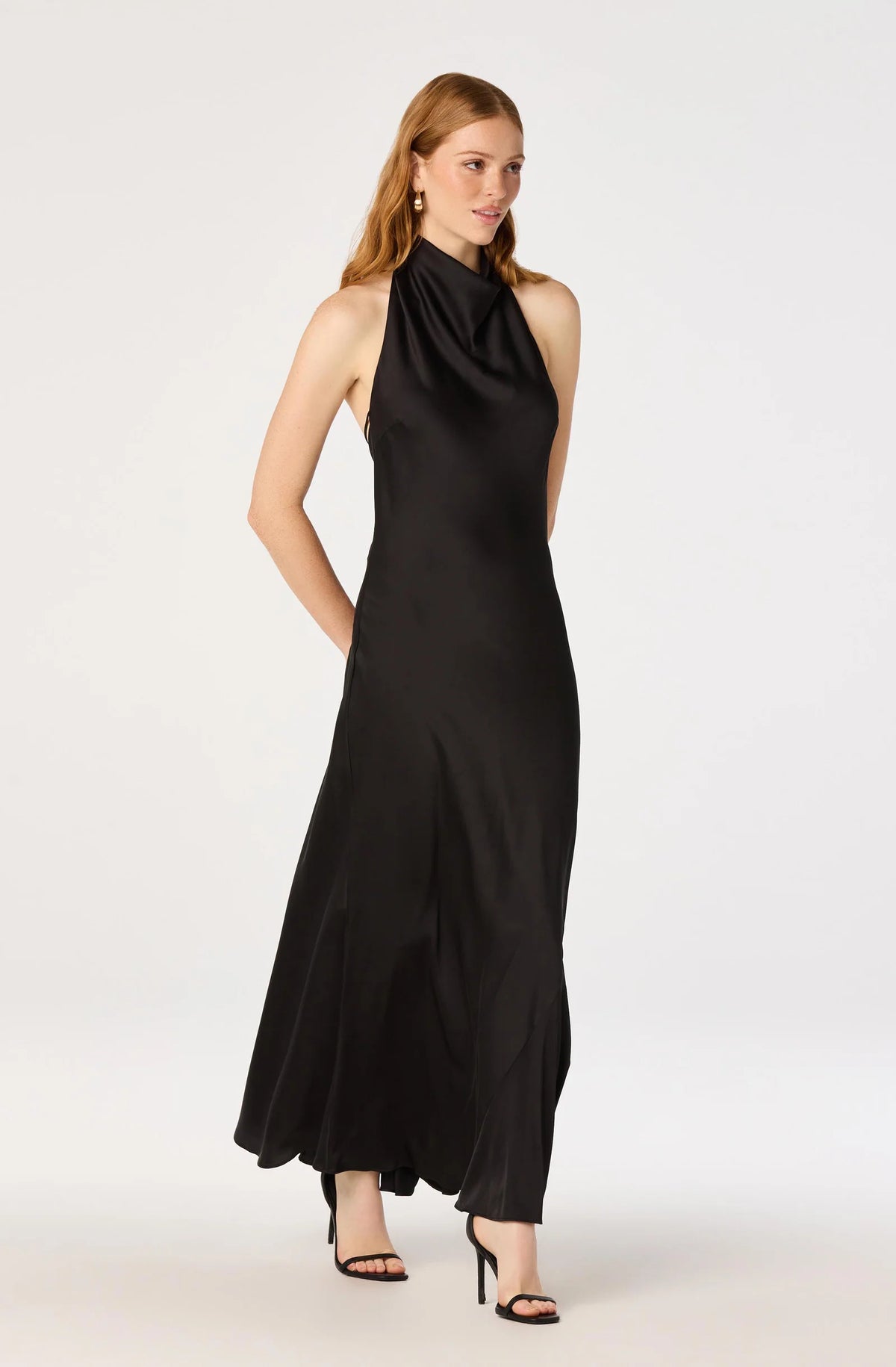 A woman modeling a sleeveless black halter-neck maxi dress with a draped satin-like finish, paired with black high-heeled sandals, against a plain light gray studio background.