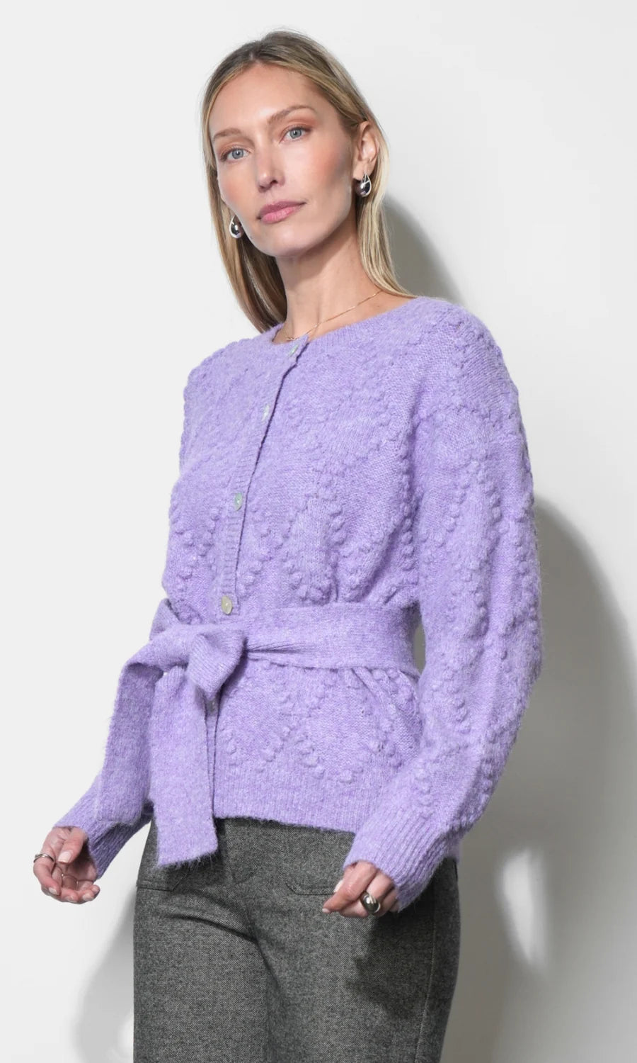 A woman modeling a lavender textured knit cardigan with a tie waist belt, paired with gray trousers, against a plain off-white wall.