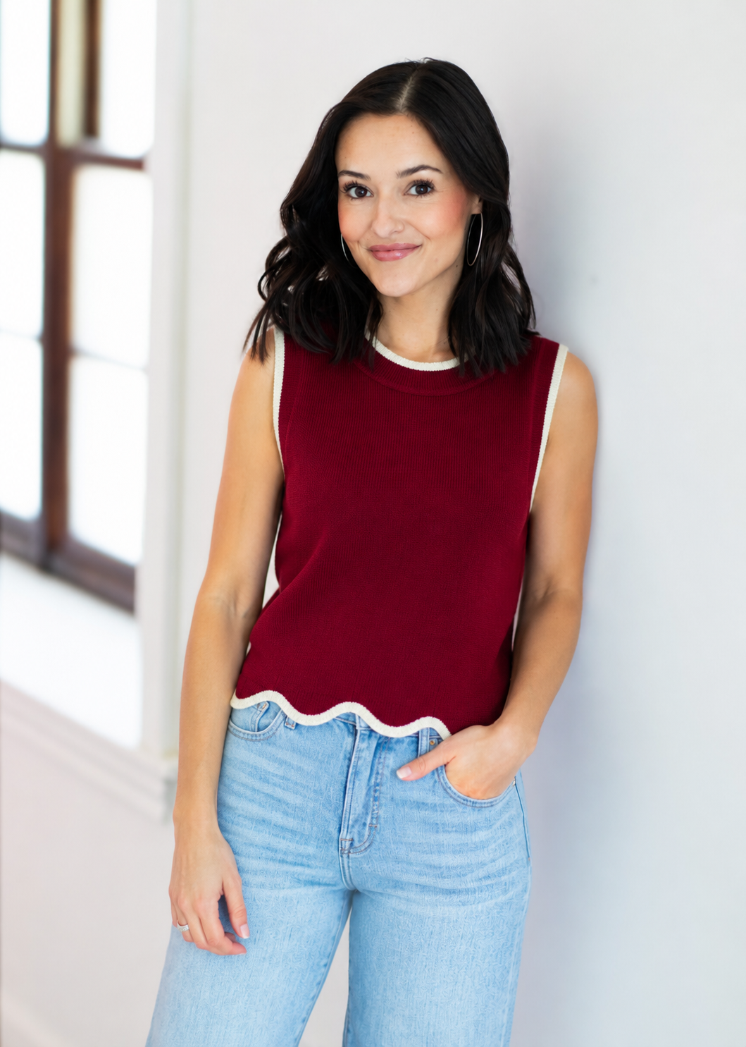 Woman wearing a red sleeveless top and light blue jeans against a white wall.
