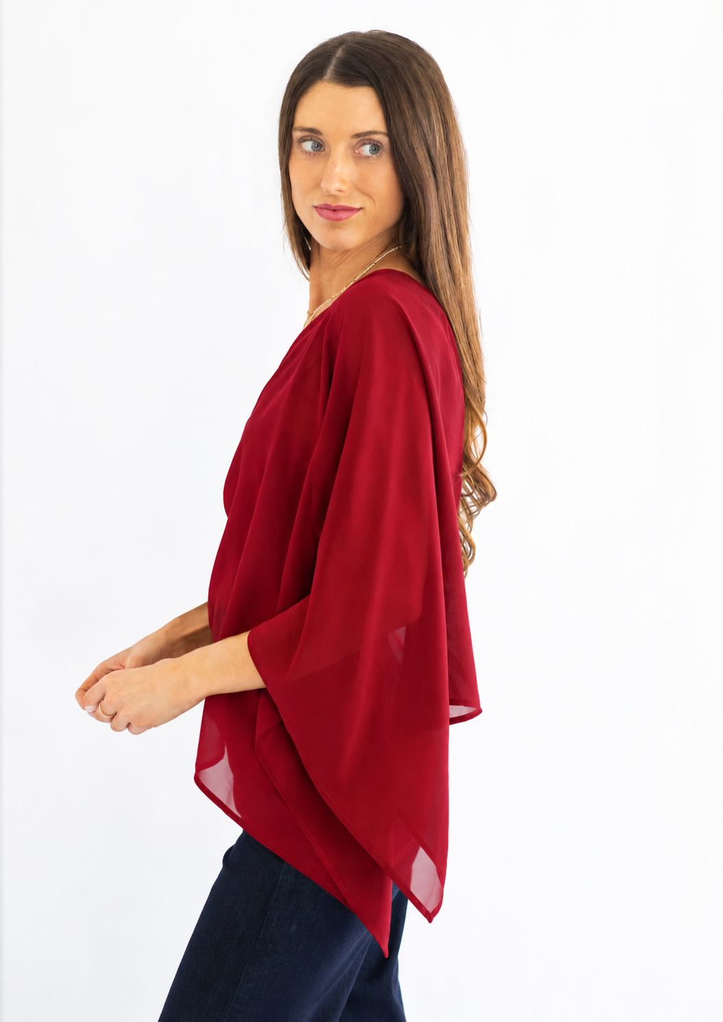 A woman wearing a red draped, semi-sheer three-quarter sleeve top paired with dark blue pants, photographed against a solid white background.