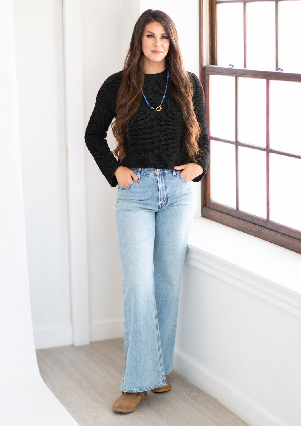 A woman stands in a bright room next to a window, wearing a black long-sleeve top, light-wash blue flare jeans, and brown shoes.