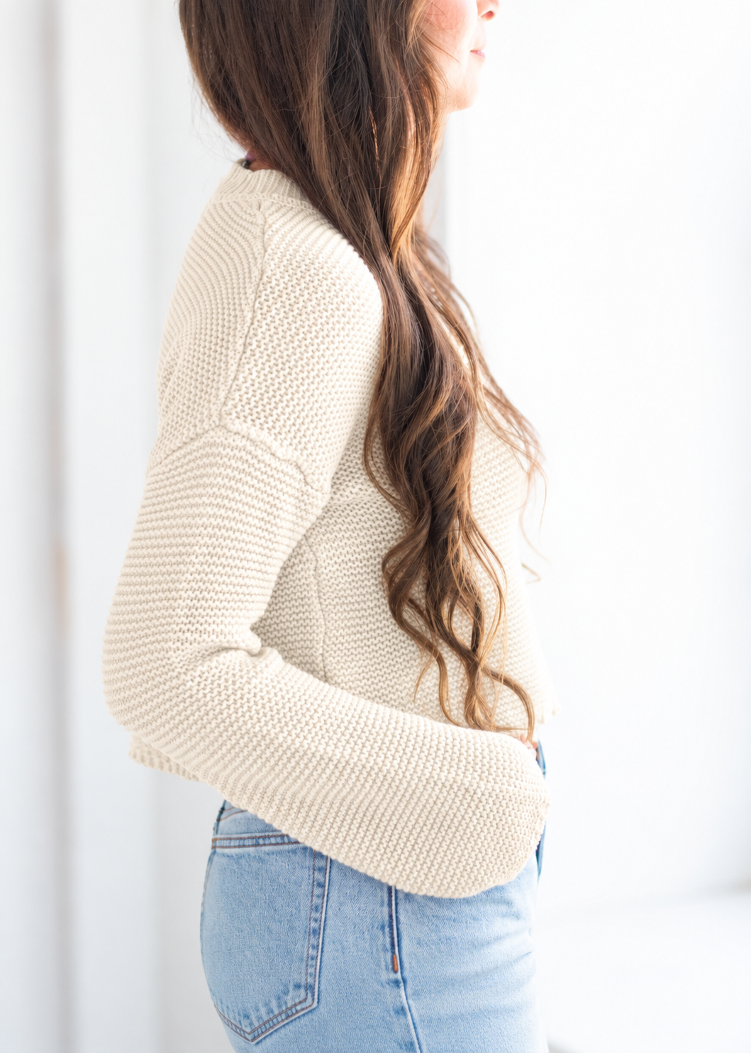 A woman wearing a cream-colored, textured knit sweater and light blue jeans, shown from a side profile against a bright white background.