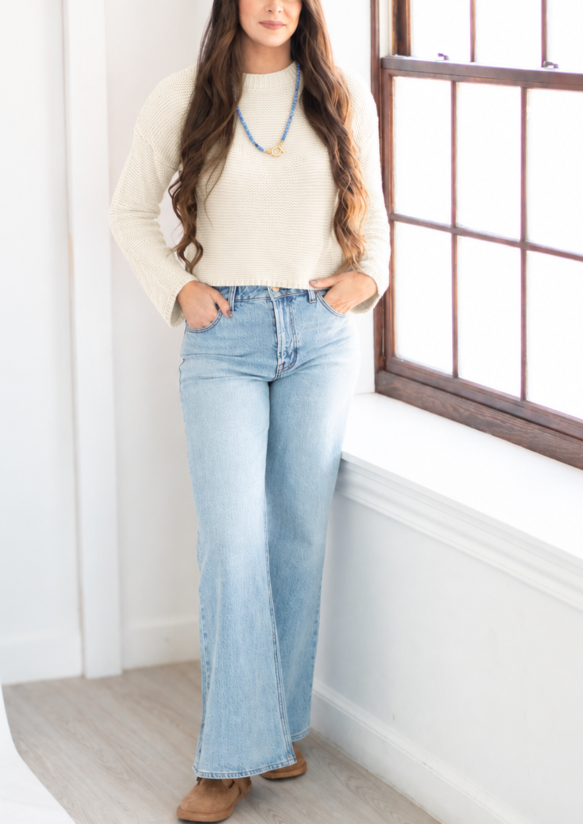 A woman stands in a bright room next to a window, wearing a cream long-sleeve knit top, light-wash blue flare jeans, and brown shoes.
