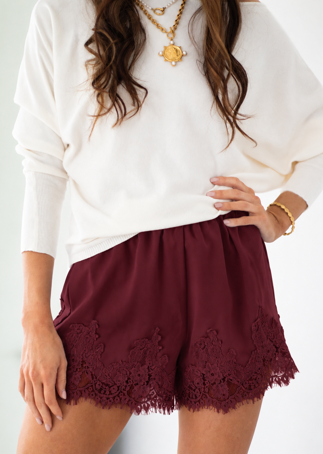 A woman wearing burgundy lace-trimmed shorts and a white long-sleeve top, accessorized with gold necklaces and bracelets, against a plain white background.