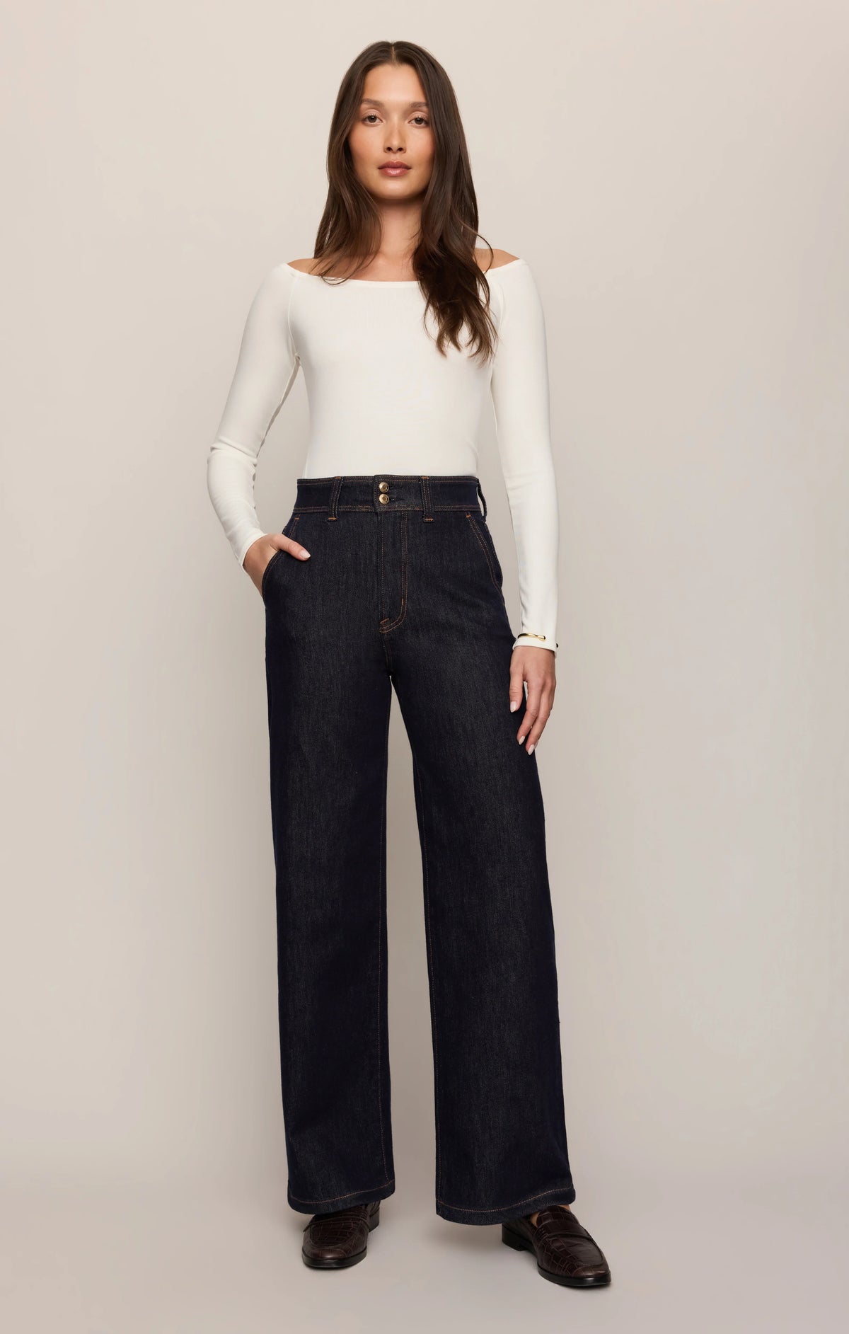 A woman modeling a pair of dark wash wide-leg jeans with a white off-the-shoulder long-sleeve top and dark brown shoes, standing against a plain light beige studio background.