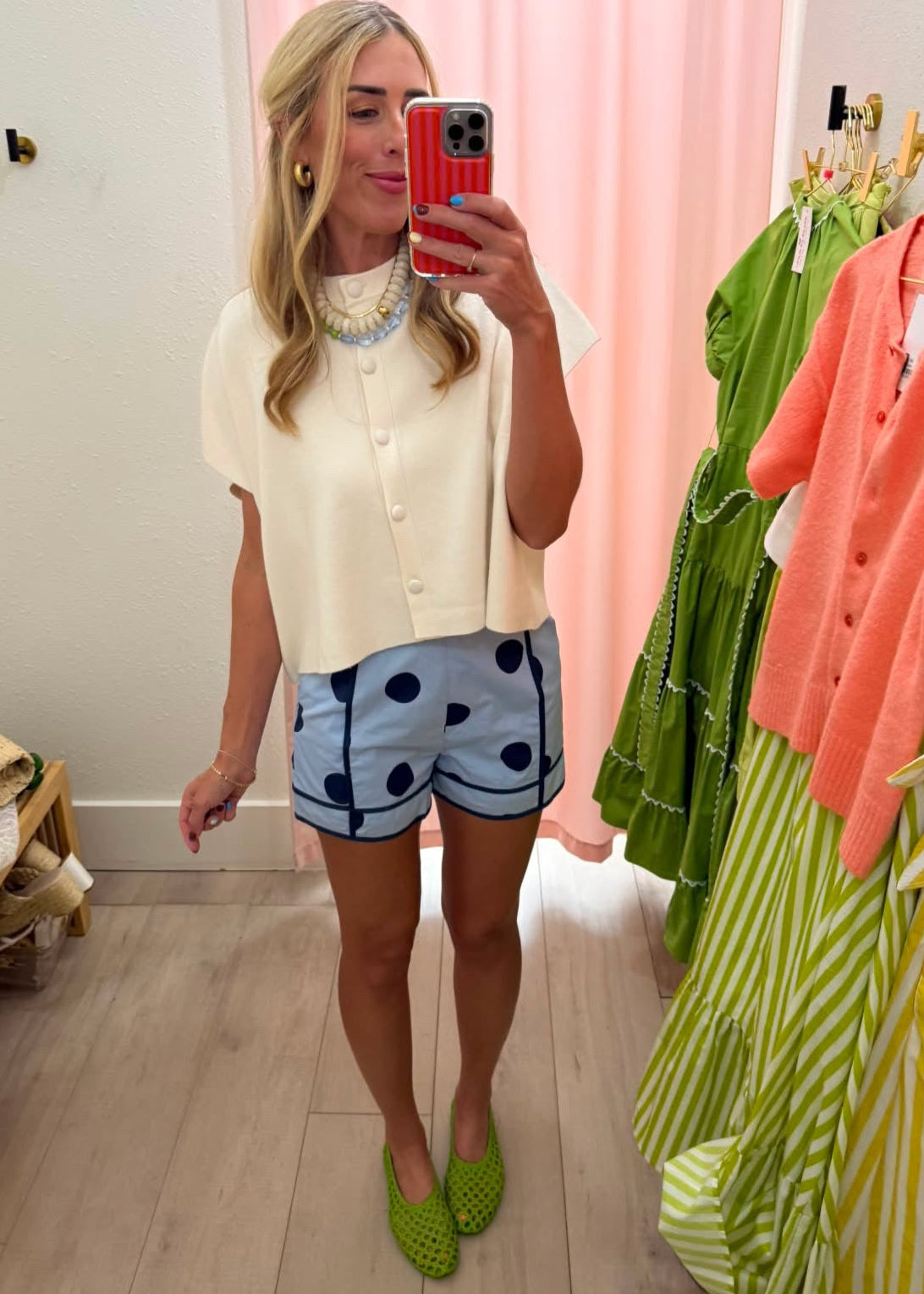 A woman takes a mirror selfie in a boutique showing off a cream short-sleeve button-up top and light blue shorts with dark blue polka dots. She is wearing bright green clogs, with clothing racks and a pink curtain visible in the background.