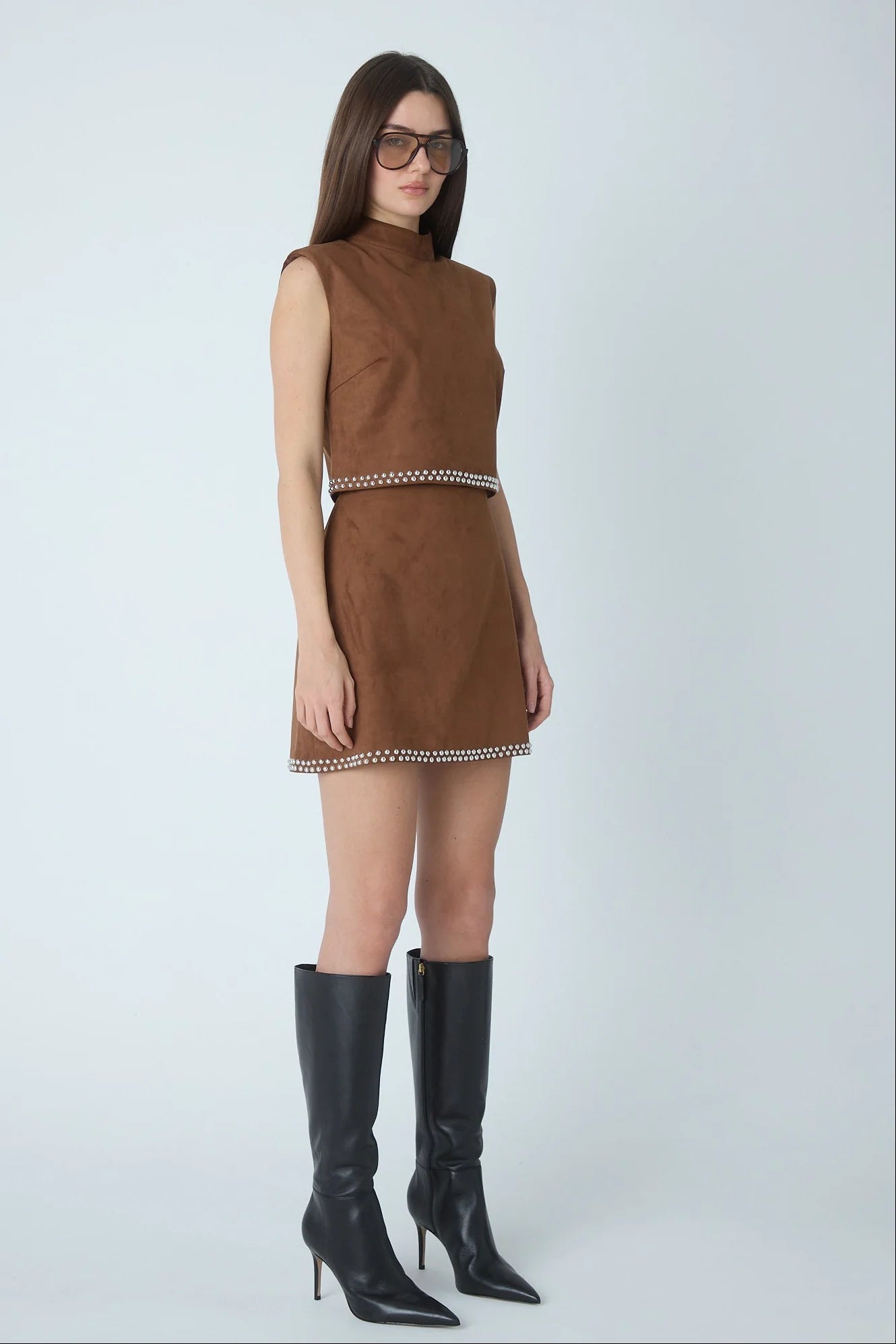A woman modeling a sleeveless brown suede matching set with a silver chain-stitched waist and hem, paired with black knee-high leather boots, against a light gray studio background.