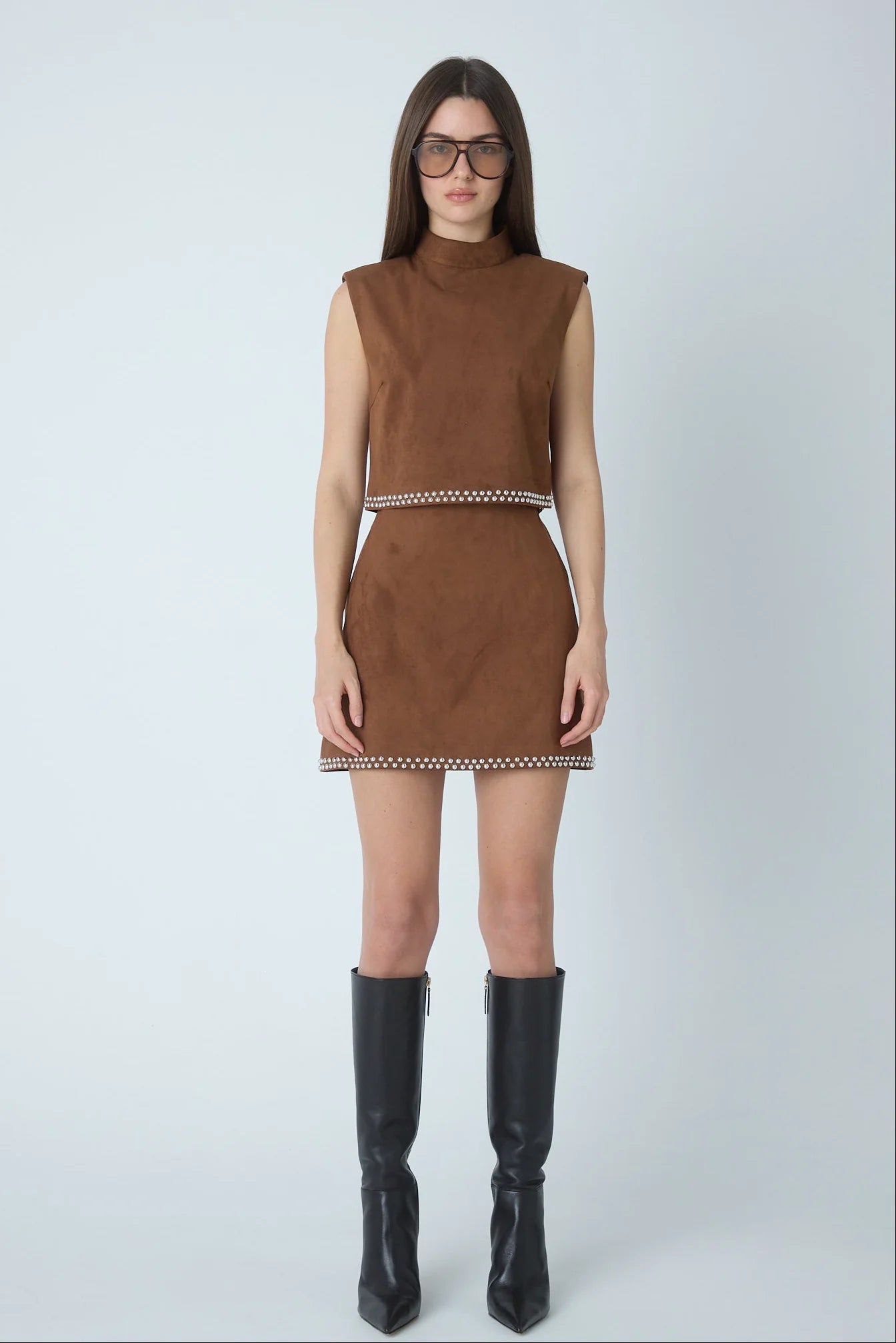 A model wearing a sleeveless brown faux-suede matching set with a mock neck and a thin black and white patterned waistband, paired with black knee-high boots against a light gray studio background.