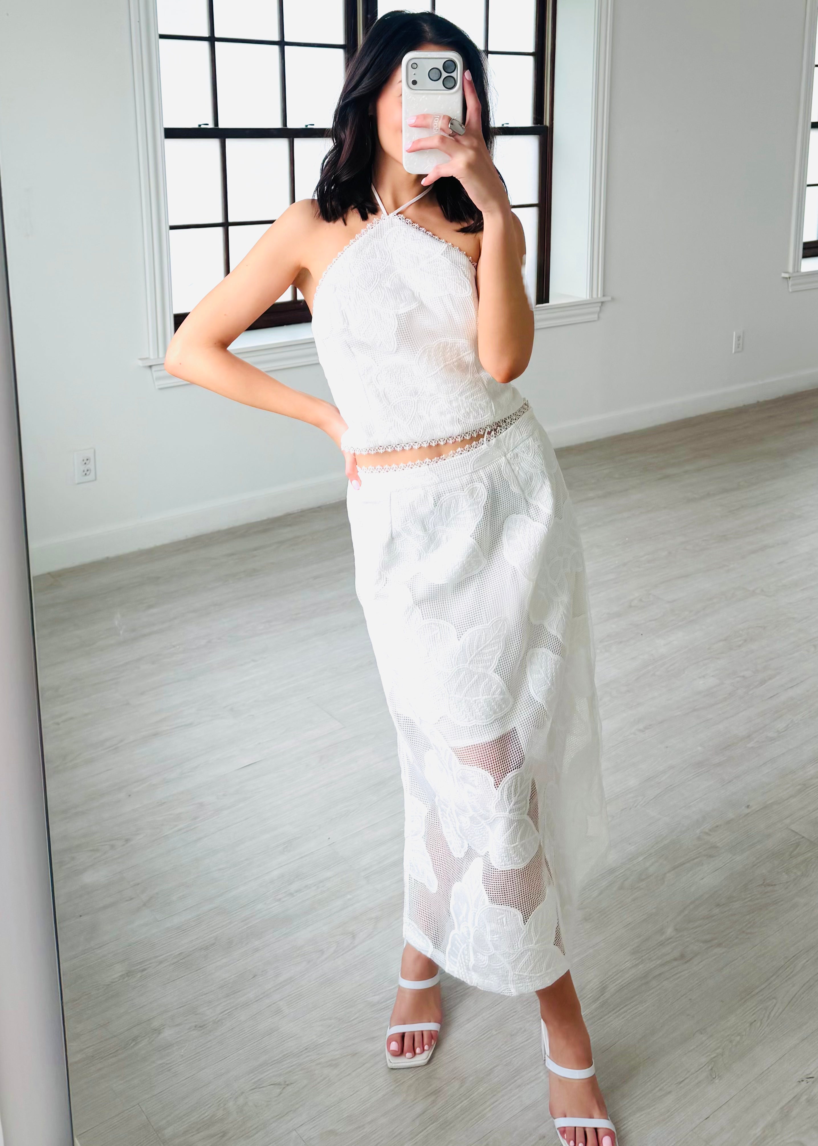 Woman taking a mirror selfie wearing a white lace outfit in a minimalistic room.