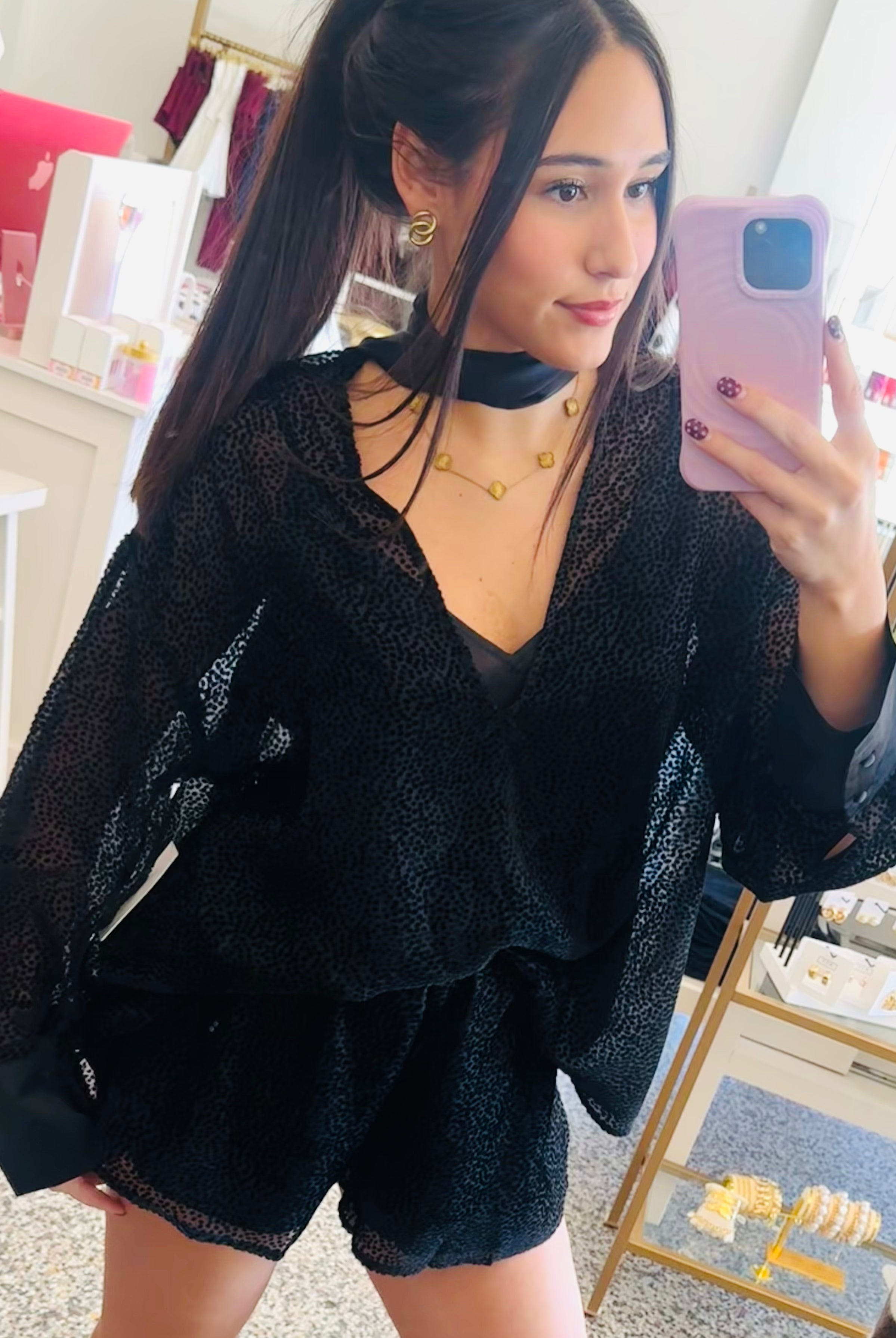 A woman takes a mirror selfie in a boutique, wearing a black sheer long-sleeve top and matching black shorts, paired with beige cowboy boots.
