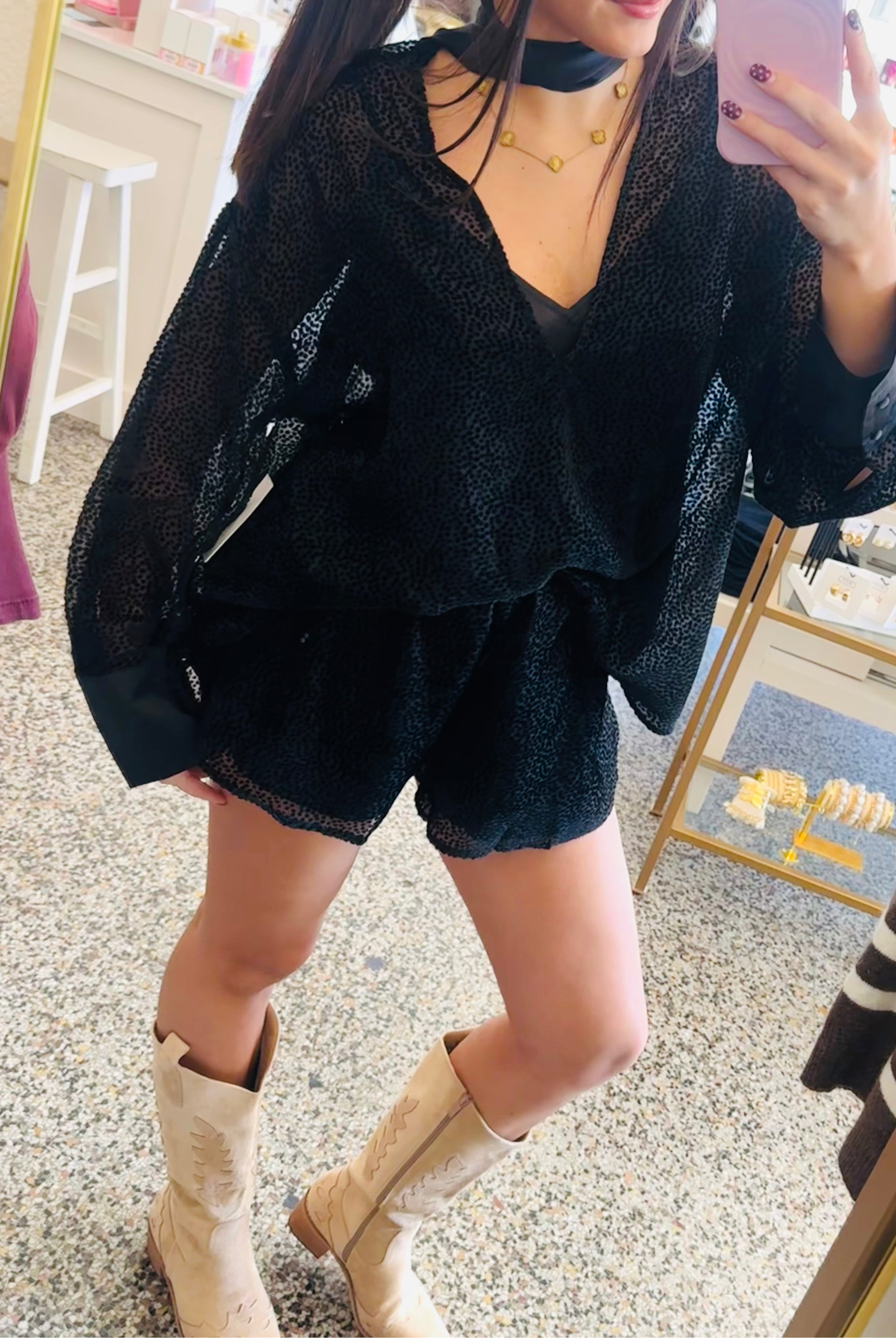 A woman takes a mirror selfie in a boutique, wearing a black sheer long-sleeve top and matching black shorts, paired with beige cowboy boots.