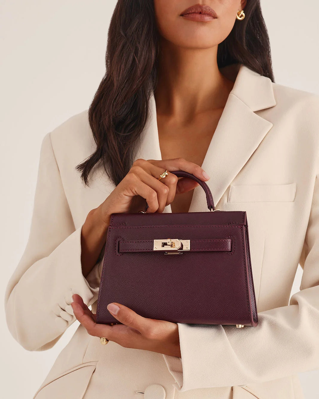 A woman in a cream blazer holds a structured top-handle handbag in deep burgundy with gold-tone hardware against a clean off-white studio background.