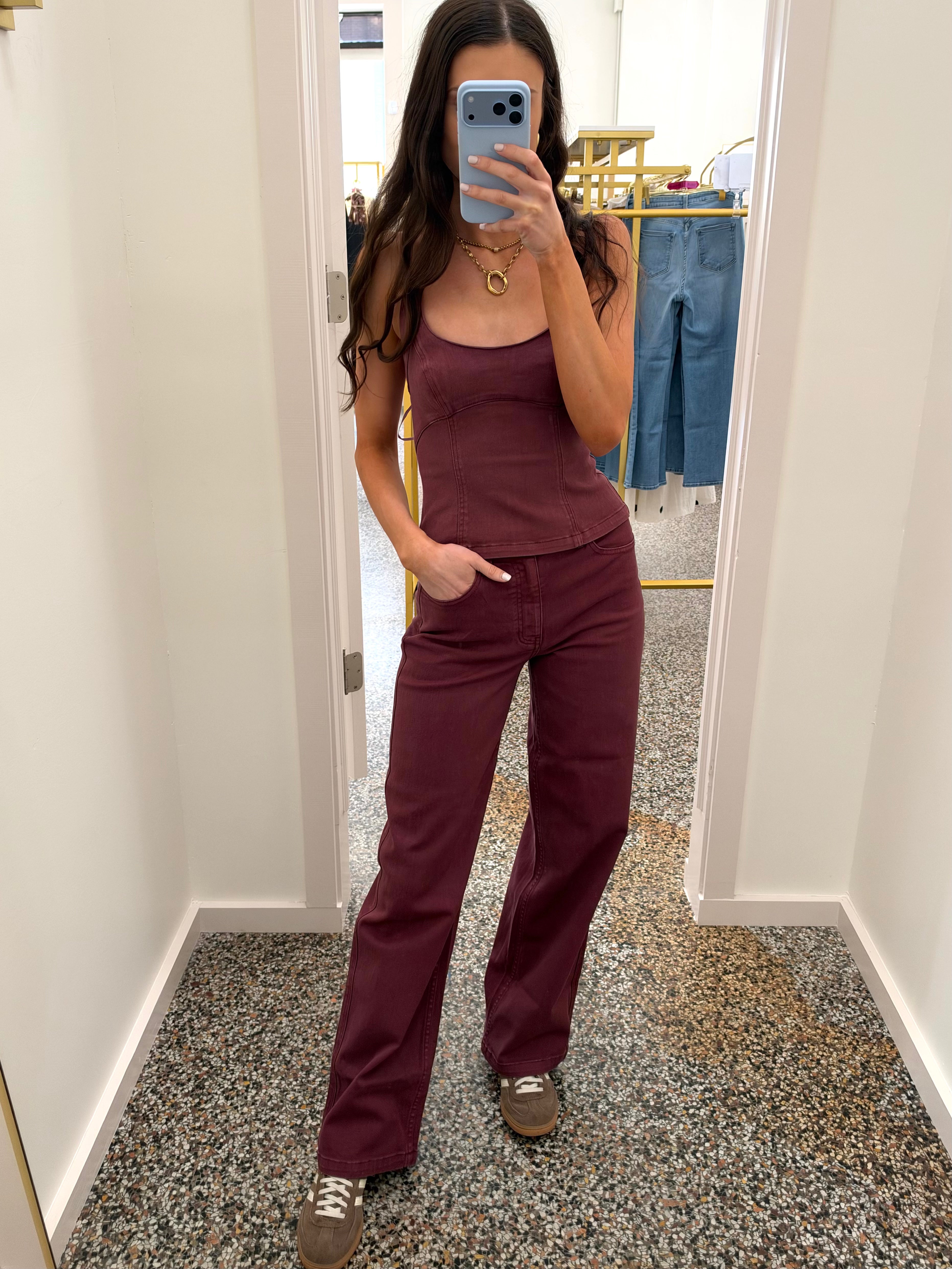 A woman takes a mirror selfie wearing a matching two-piece set consisting of a sleeveless scoop-neck camisole tank top and wide-leg trousers, both in a deep burgundy color. She is standing in a fitting room with a speckled floor and a clothing rack visible in the background.