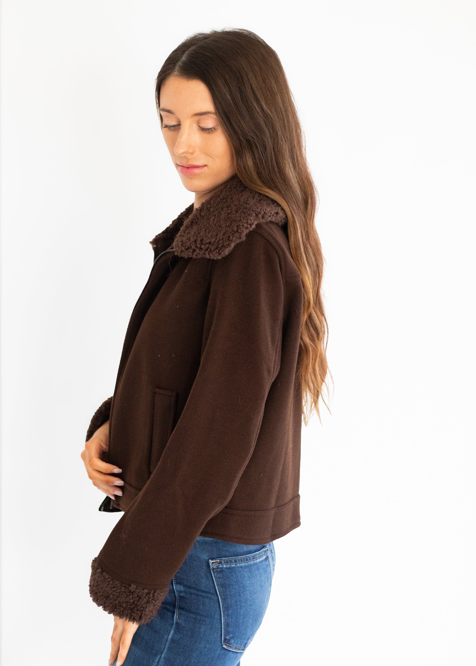 A woman wearing a dark brown suede-like jacket with a brown faux-shearling collar and cuffs, paired with blue jeans, photographed against a plain white background.