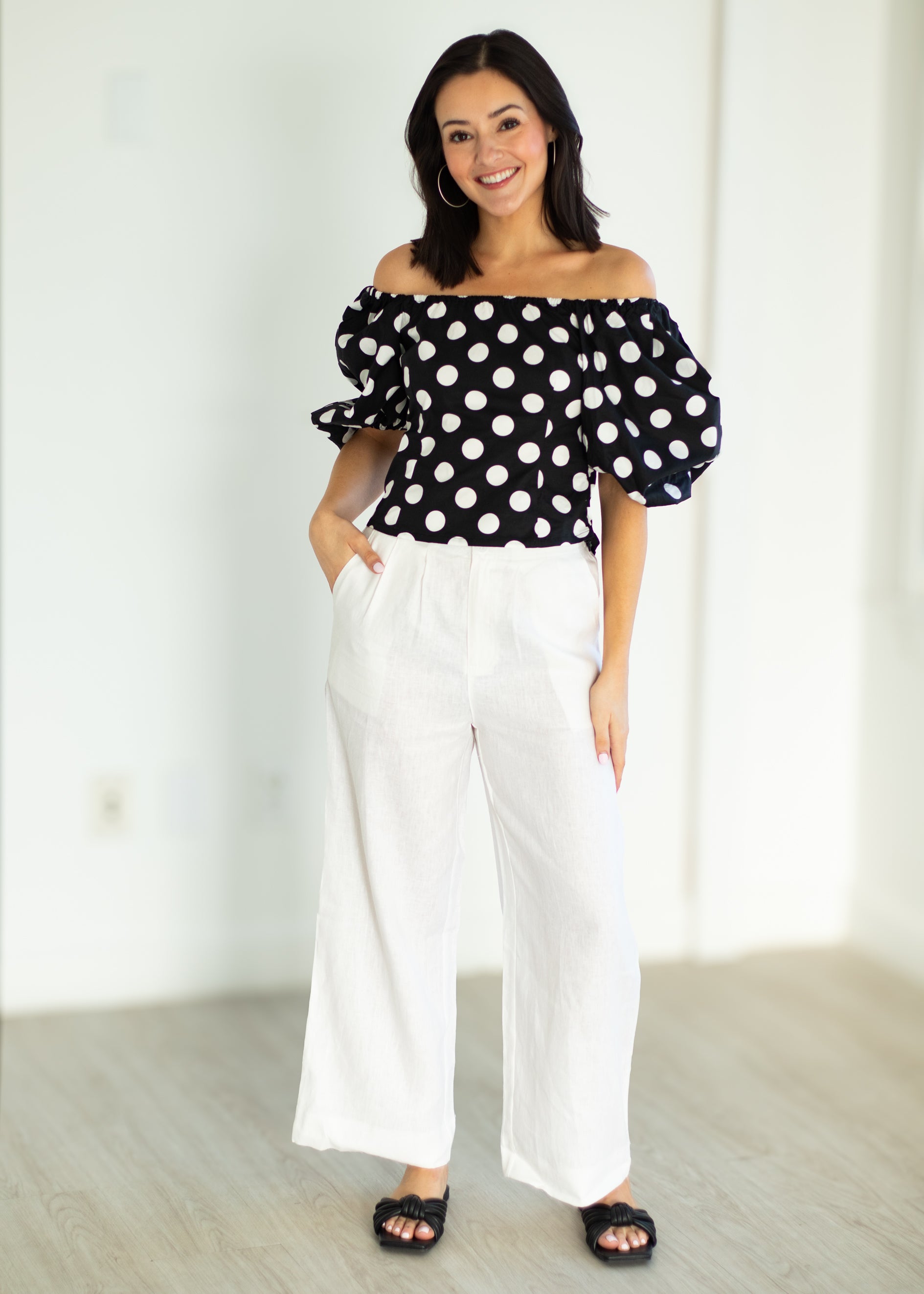 Woman wearing a black polka dot top and white pants in a minimal indoor setting