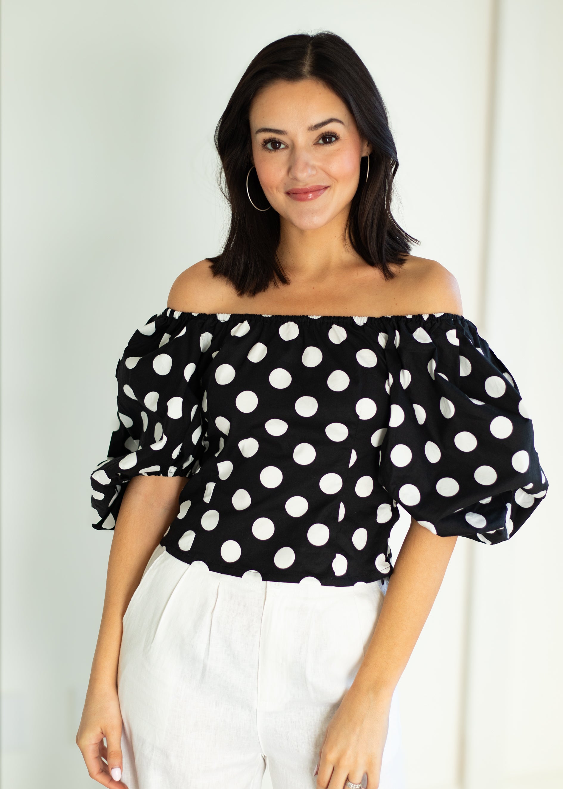 Woman wearing a black off-shoulder top with white polka dots and white pants against a plain background
