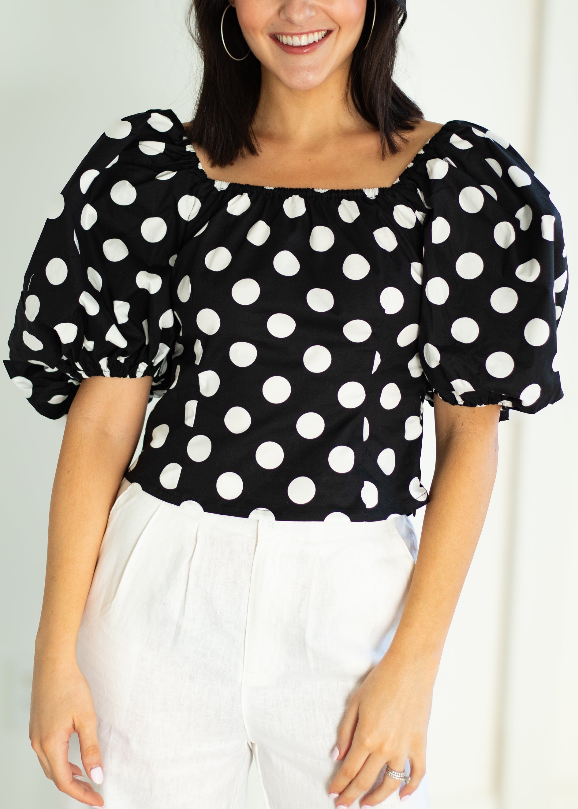Woman wearing a black polka dot blouse with white pants against a light background